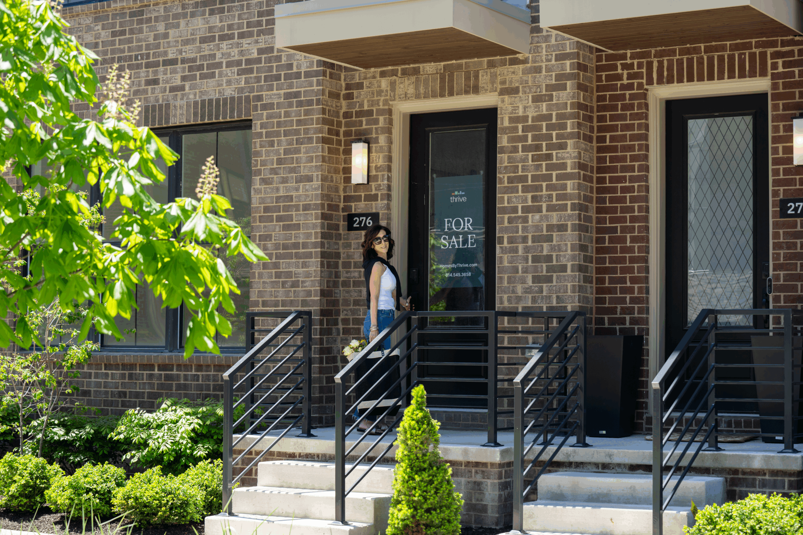 Woman standing at the door of a brick townhouse with a For Sale sign, surrounded by greenery and steps leading to the entrance.
