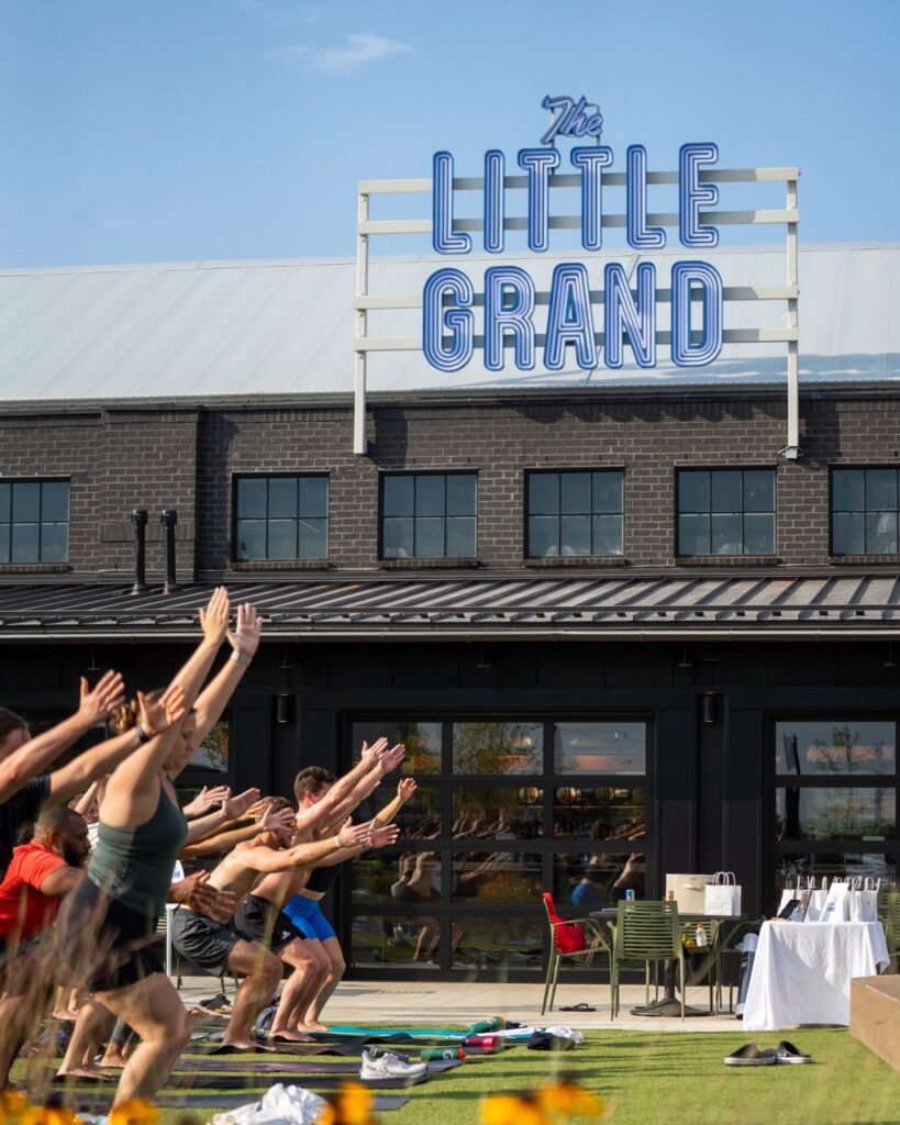 People participating in an outdoor yoga class in front of a building with a sign reading The Little Grand.