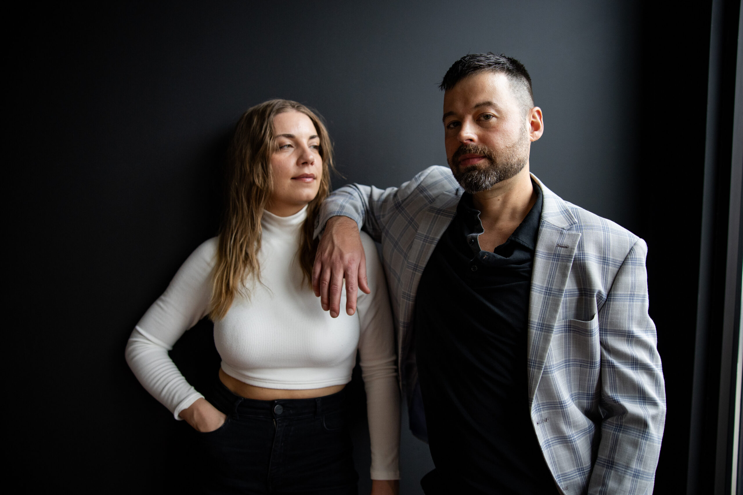 Thrive Companies, A woman in a white turtleneck top stands next to a man in a plaid blazer and black shirt, who is leaning with his arm on her shoulder against a dark background. Apartments in Columbus Ohio