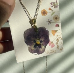 Thrive Companies, A necklace with a pressed purple flower pendant is displayed on a floral card, held up in sunlight with shadowed lines in the background. Apartments in Columbus Ohio