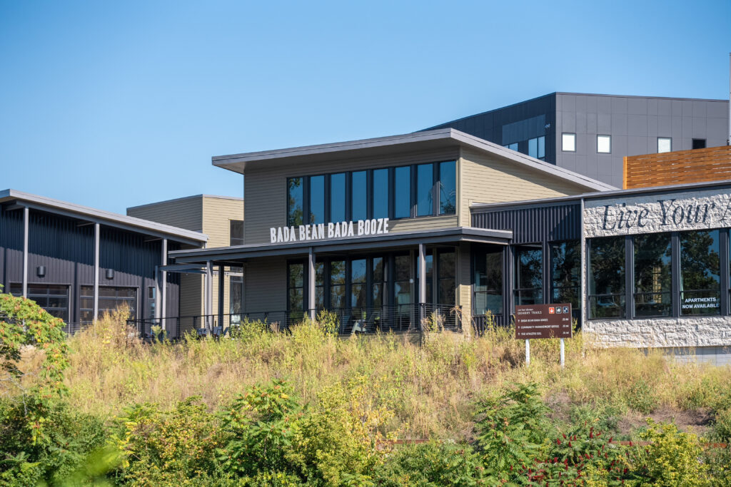 Modern commercial building with large windows and a sign reading BAD BEAN BAD BOOZE, surrounded by overgrown plants and greenery under a clear sky.