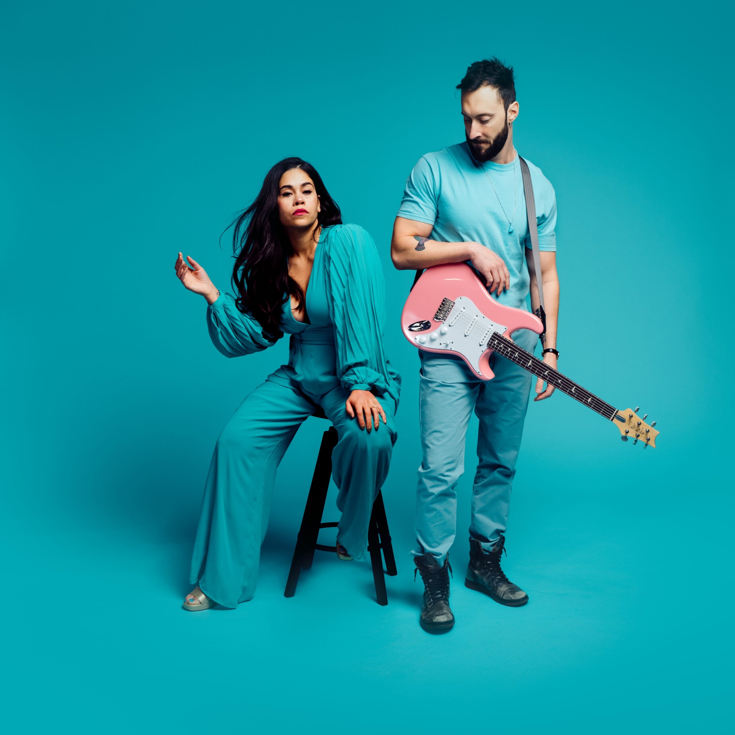 Thrive Companies, A woman in a teal jumpsuit sits on a stool while a man in teal clothes stands holding a pink electric guitar, both posing against a matching teal background. Apartments in Columbus Ohio