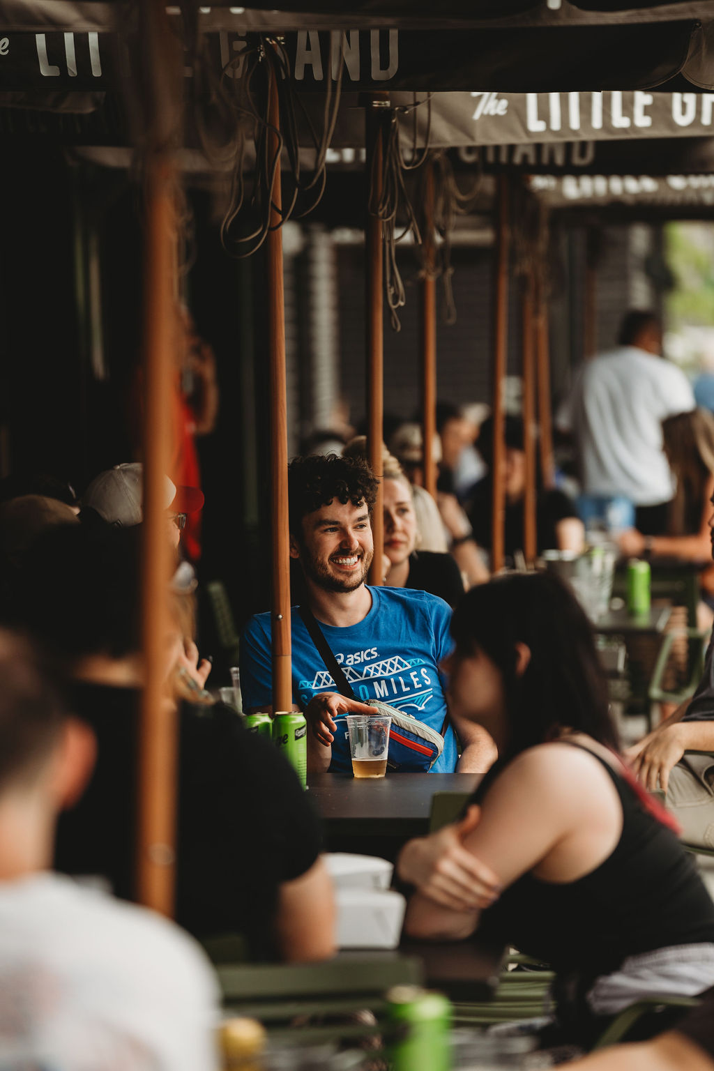 Thrive Companies, A man in a blue shirt smiles while sitting at an outdoor table with drinks, surrounded by other people at a busy cafe or restaurant. Apartments in Columbus Ohio