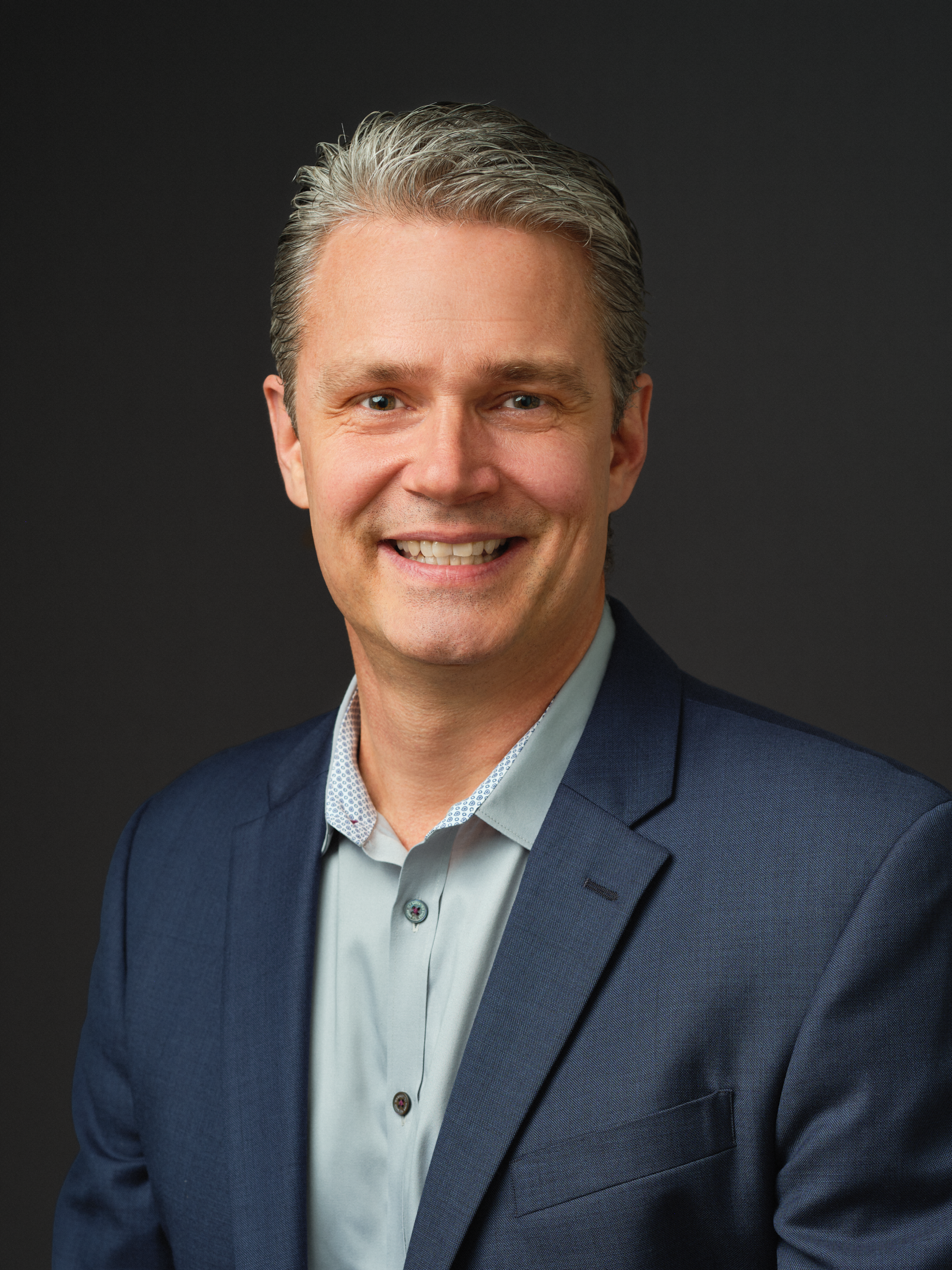 Thrive Companies, A middle-aged man with short gray hair, wearing a blue blazer and light-colored shirt, smiles in a professional studio portrait with a plain dark background. Apartments in Columbus Ohio