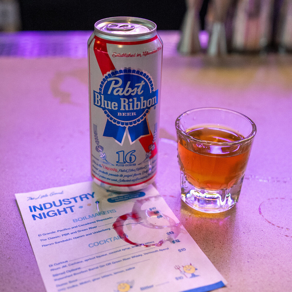 Thrive Companies, A can of Pabst Blue Ribbon beer, a shot of whiskey, and a drink menu labeled Industry Night sit on a bar counter with drink rings. Apartments in Columbus Ohio