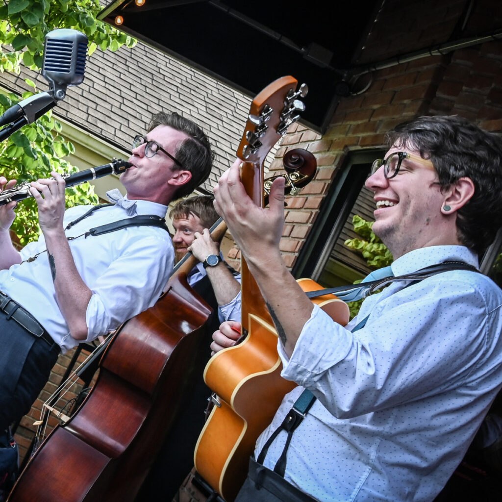 Thrive Companies, Three musicians play instruments—clarinet, double bass, and guitar—outdoors under a canopy, dressed in white shirts and suspenders, with a vintage microphone nearby. Apartments in Columbus Ohio