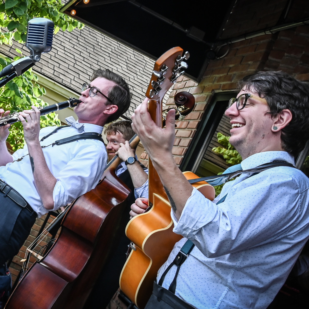 Thrive Companies, Three musicians play instruments—clarinet, double bass, and guitar—outdoors under a canopy, dressed in white shirts and suspenders, with a vintage microphone nearby. Apartments in Columbus Ohio