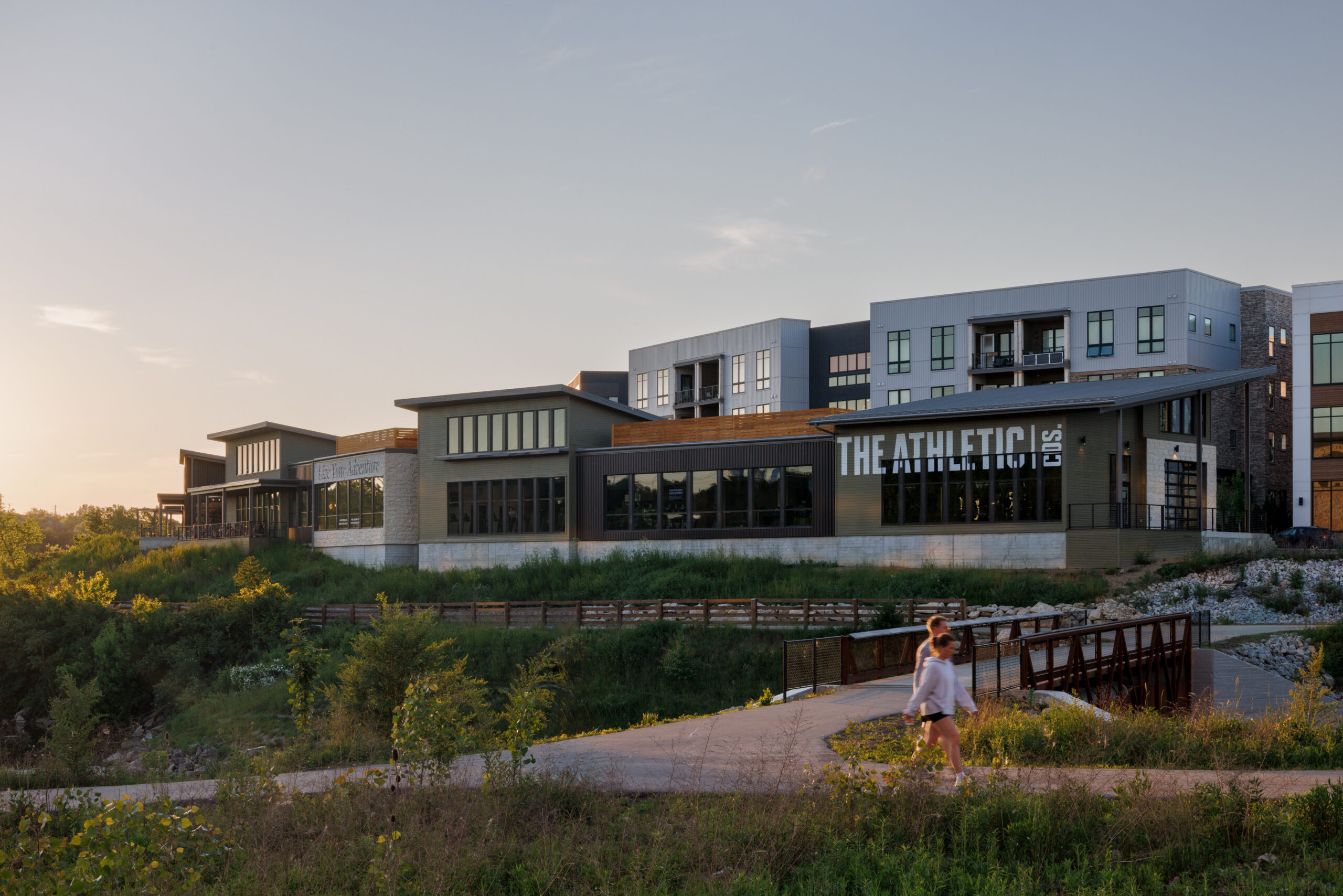 Thrive Companies, A modern building with THE ATHLETIC signage is set on a grassy landscape at sunset, with a person walking on a nearby path and bridge in the foreground. Apartments in Columbus Ohio