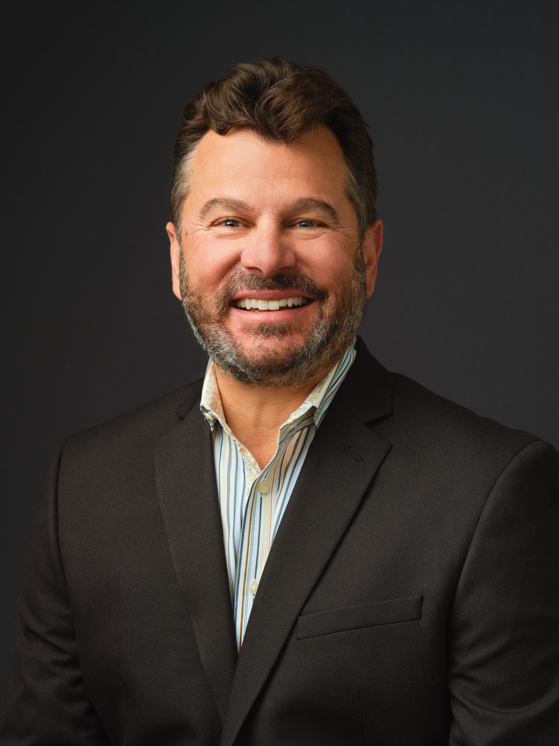 Thrive Companies, A man with short brown hair and a beard, wearing a dark suit jacket and striped shirt, smiles in front of a dark background. Apartments in Columbus Ohio