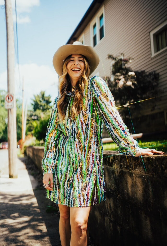 Thrive Companies, A person wearing a wide-brimmed hat and a colorful, sequined dress stands and smiles on a sunny sidewalk next to a residential building. Apartments in Columbus Ohio