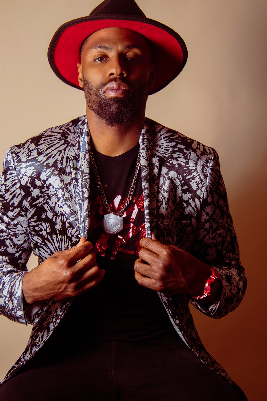 Thrive Companies, A man wearing a patterned blazer, black shirt, red and black hat, and jewelry sits against a plain background, holding the lapels of his jacket. Apartments in Columbus Ohio