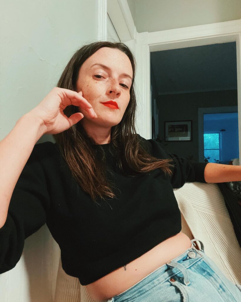 Thrive Companies, A woman with long brown hair, wearing a black crop top and jeans, sits indoors with one hand resting on her face and a neutral expression. Apartments in Columbus Ohio