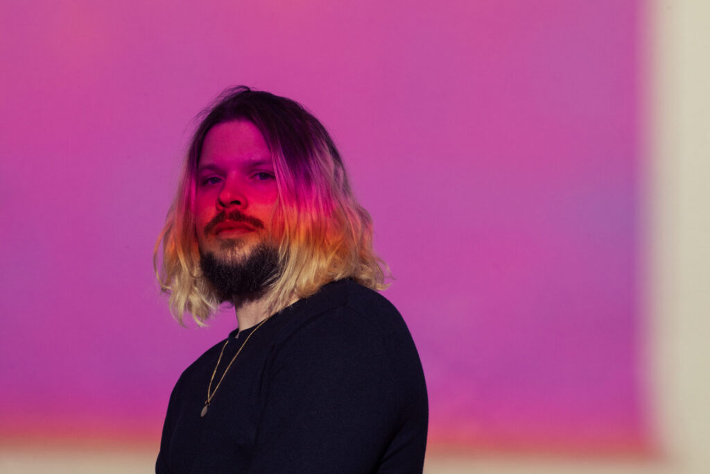 Thrive Companies, A person with long blonde hair and a beard stands against a pink and purple gradient background, wearing a dark shirt and a necklace. Apartments in Columbus Ohio