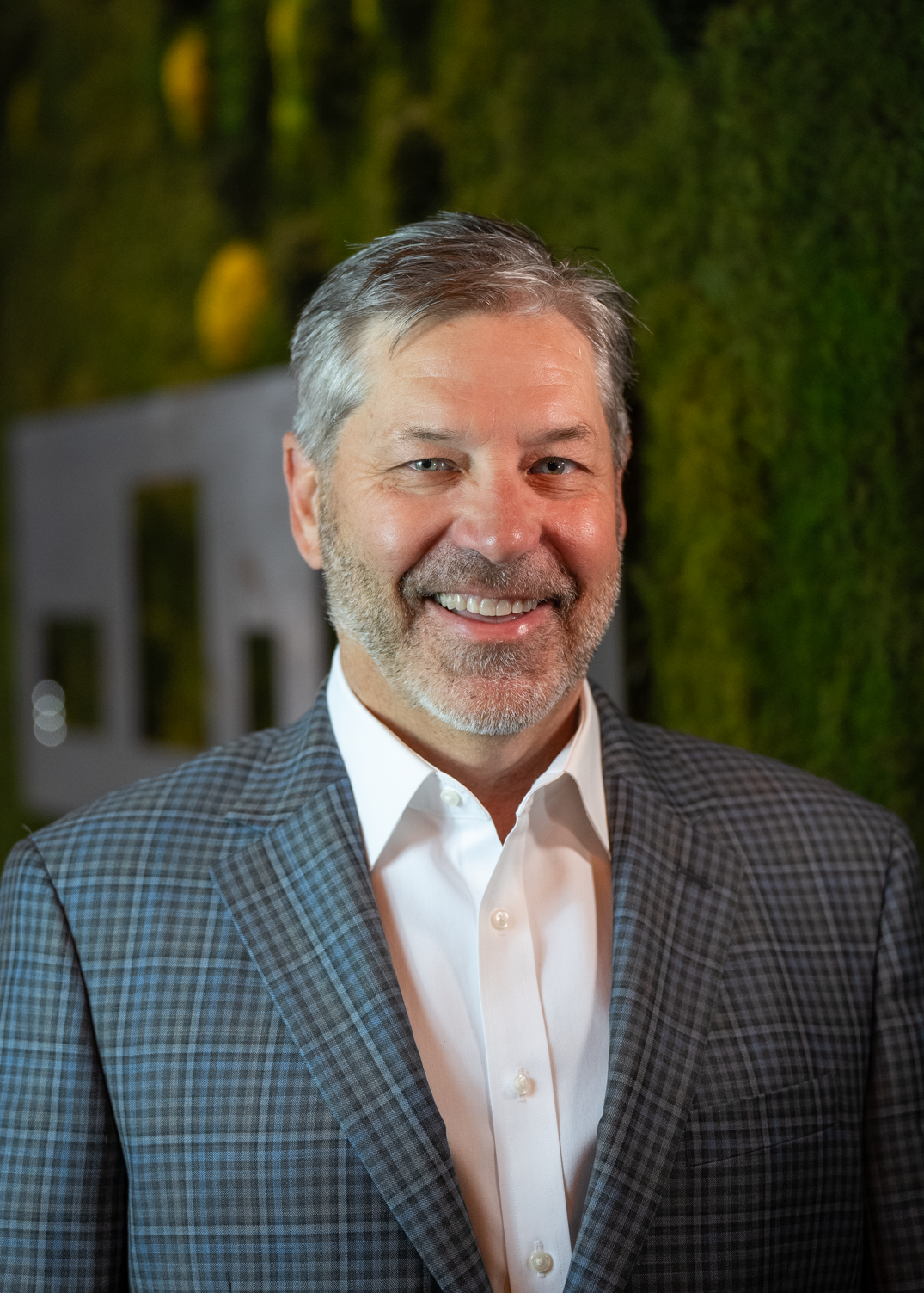 A man with gray hair and a beard, wearing a plaid suit jacket and white shirt, stands smiling in front of a blurred green background, representing the professionalism of Our Team.