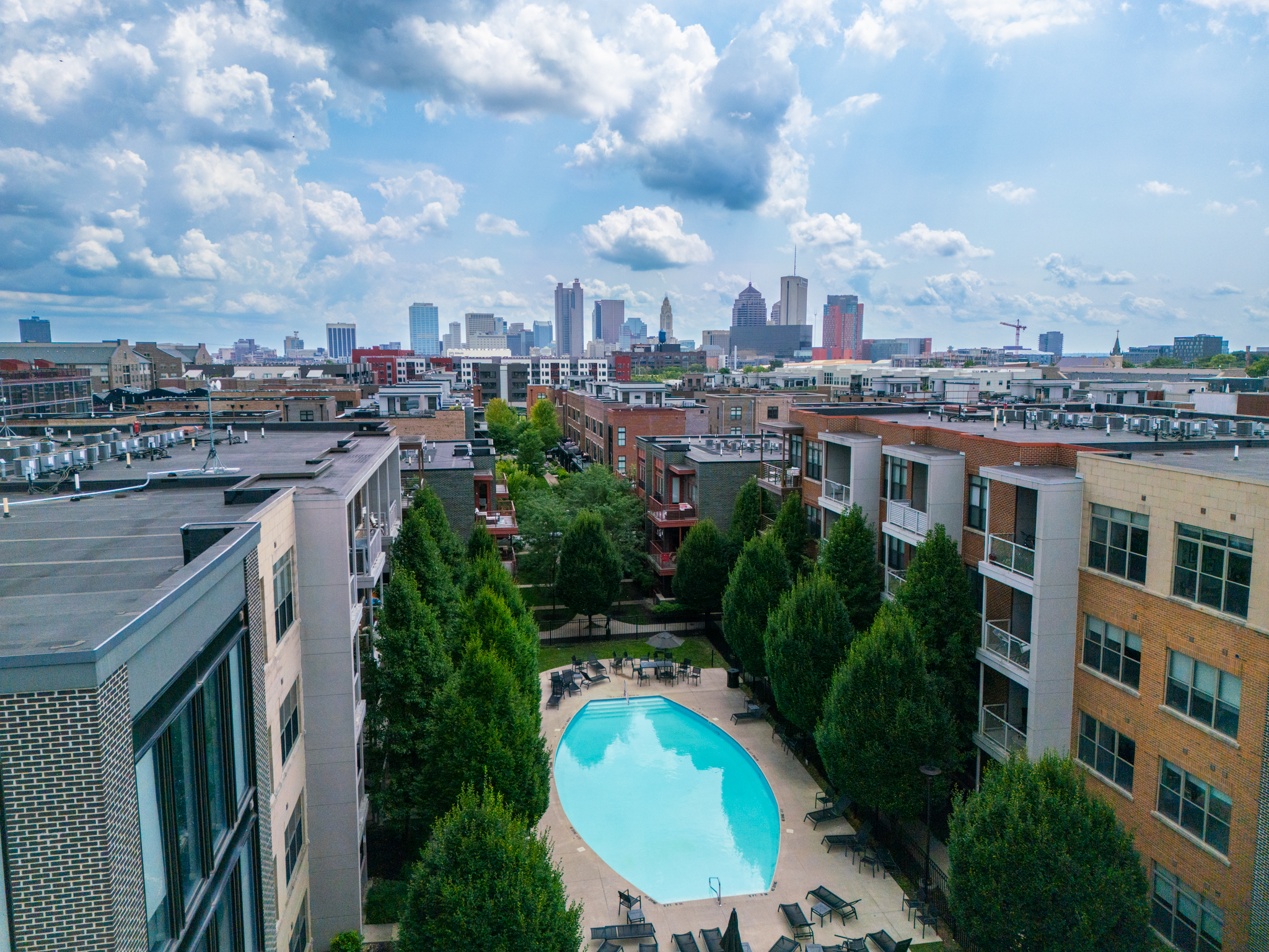 Aerial view of an apartment complex with a central swimming pool, lush trees, and nearby buildings—enjoy city skyline views and ask about our Preferred Employer Program for exclusive benefits under the partly cloudy sky.