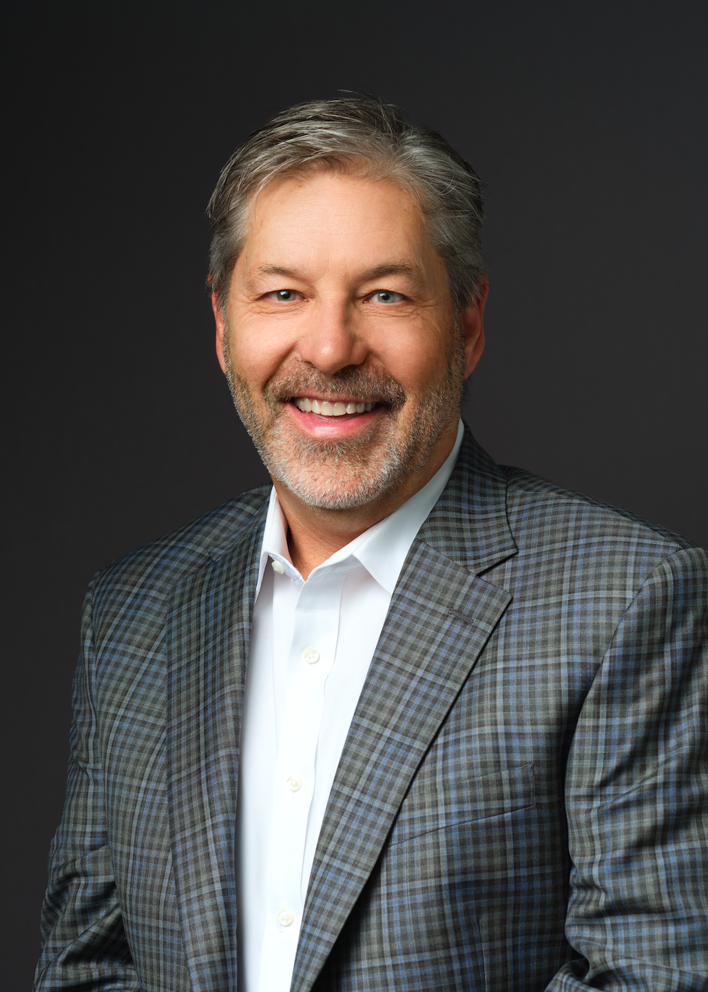 Smiling middle-aged man with gray hair and beard, wearing a checked suit jacket and white shirt, represents the dedication of Our Team, posed against a dark background.