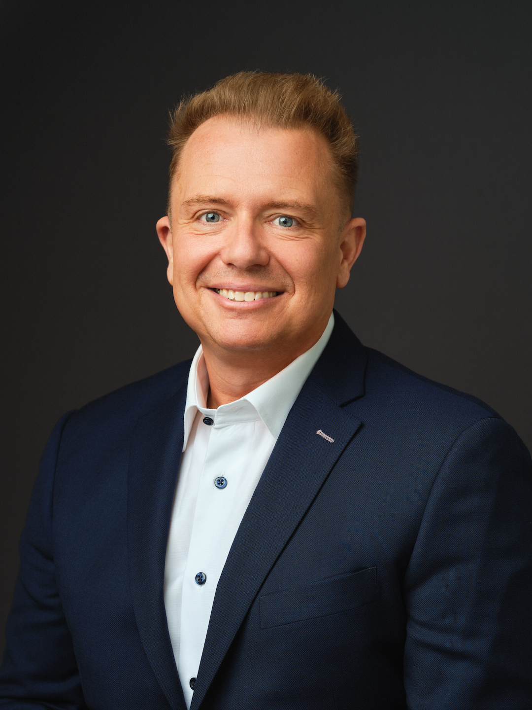A member of Our Team, a man with light hair wears a dark blue suit jacket and white dress shirt, smiling at the camera against a plain dark background.
