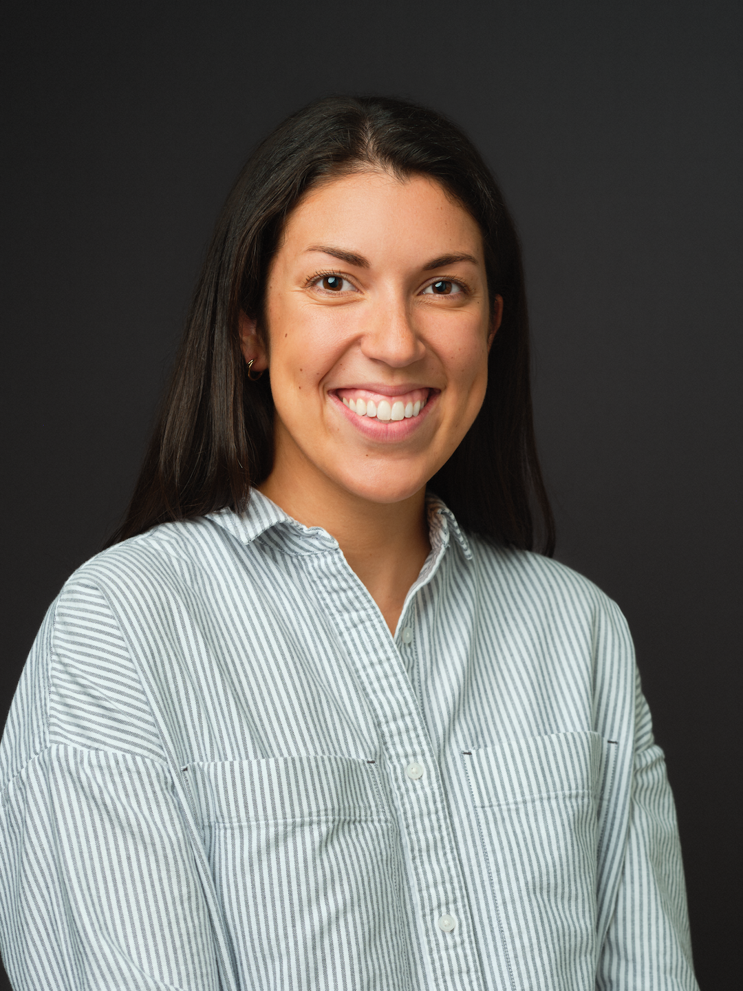 A woman with long dark hair, part of Our Team, wears a light blue and white striped button-up shirt and smiles at the camera in front of a plain dark background.