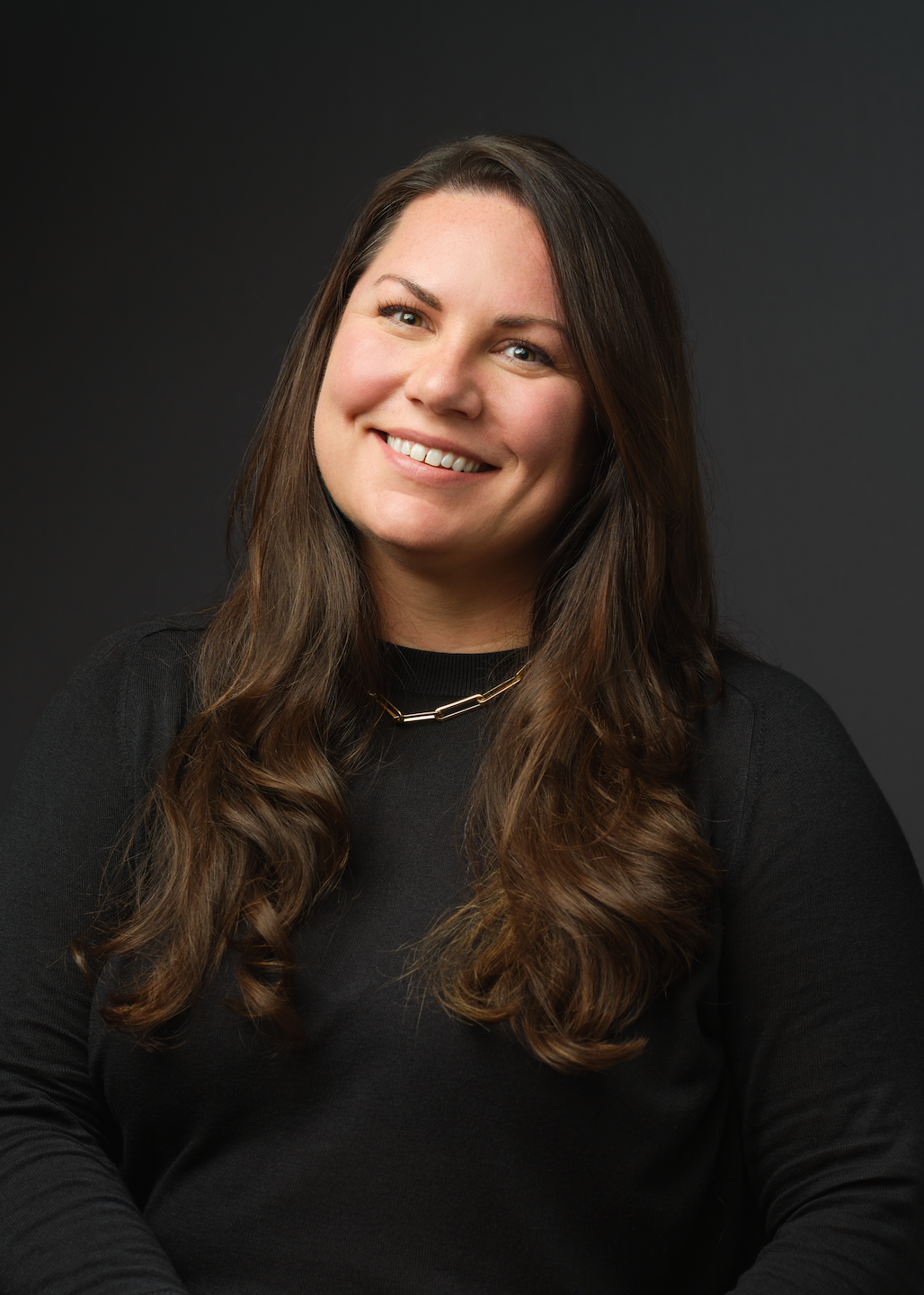 A member of Our Team, this woman with long brown hair, wearing a black top and a necklace, smiles at the camera against a dark background.