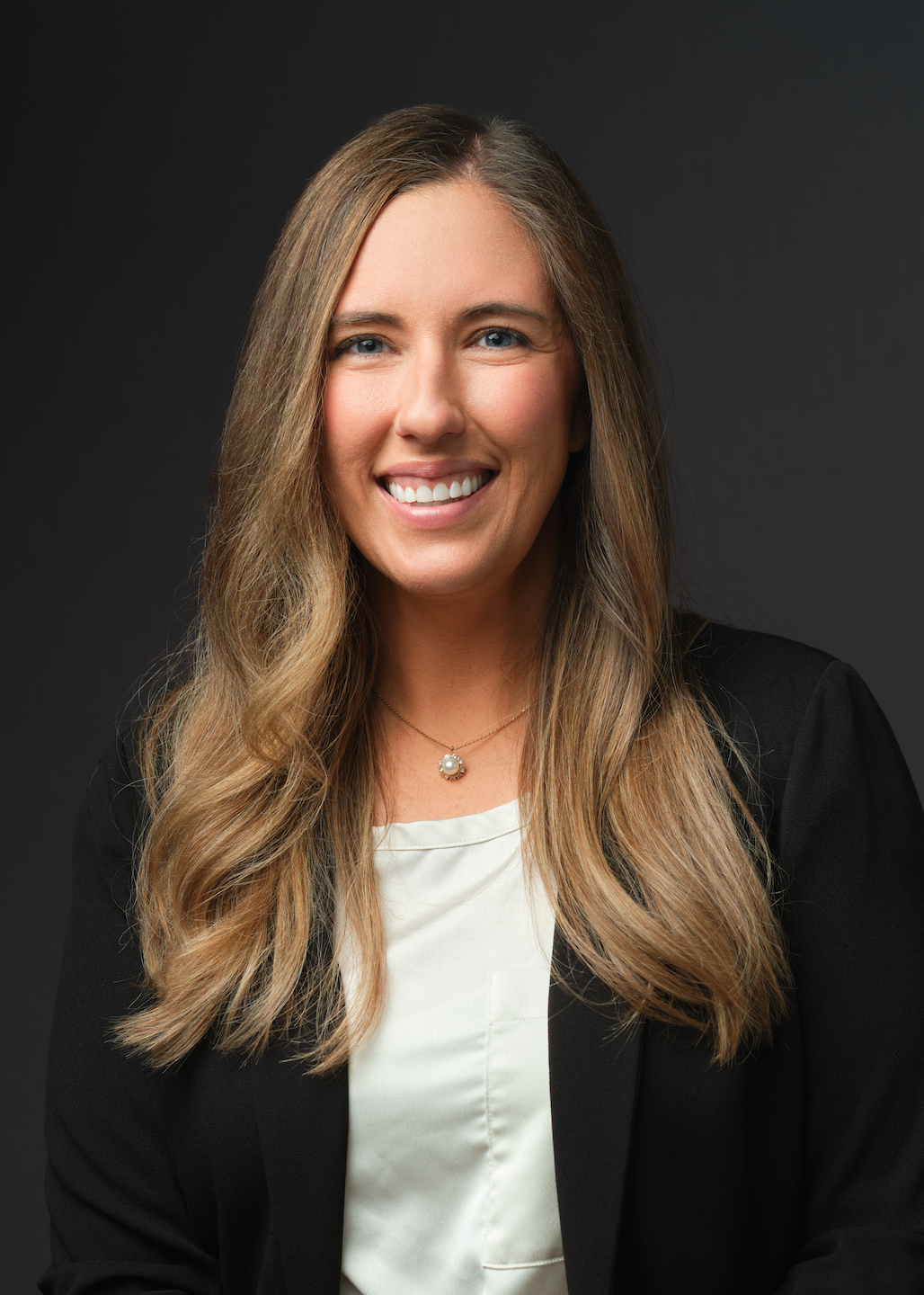 A member of Our Team, a woman with long, wavy light brown hair wearing a black blazer and white blouse, smiles in front of a dark background.