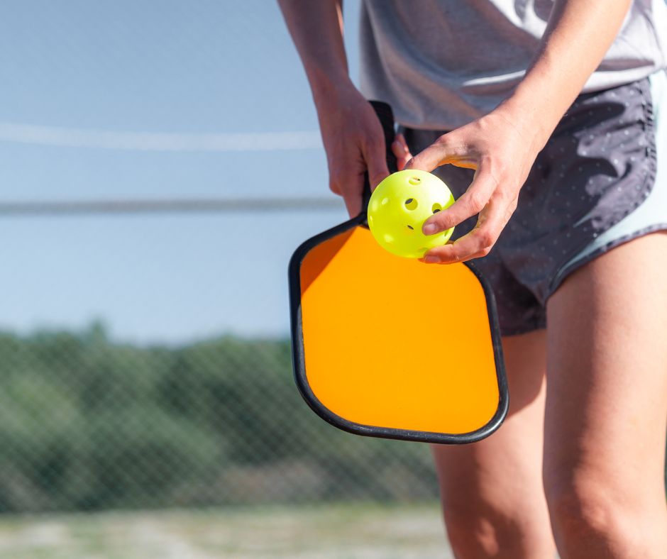 Person holding a bright orange pickleball paddle and a yellow pickleball, standing on an outdoor court at The Michael at GVX, with a blurry fence and trees in the background.