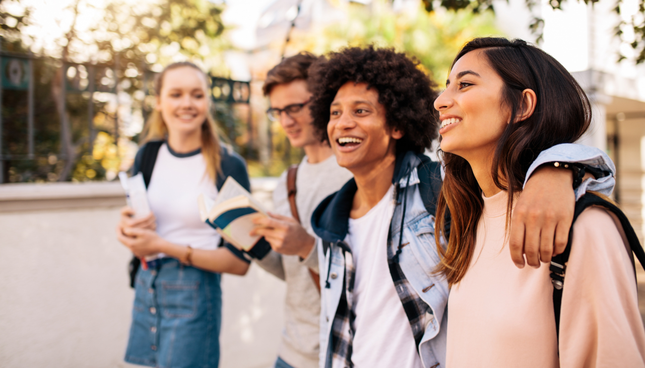 Thrive Companies, Four young adults walk outdoors, smiling and talking near student housing in Columbus. One person has their arm around another. They carry notebooks and backpacks, suggesting a school setting. Apartments in Columbus Ohio