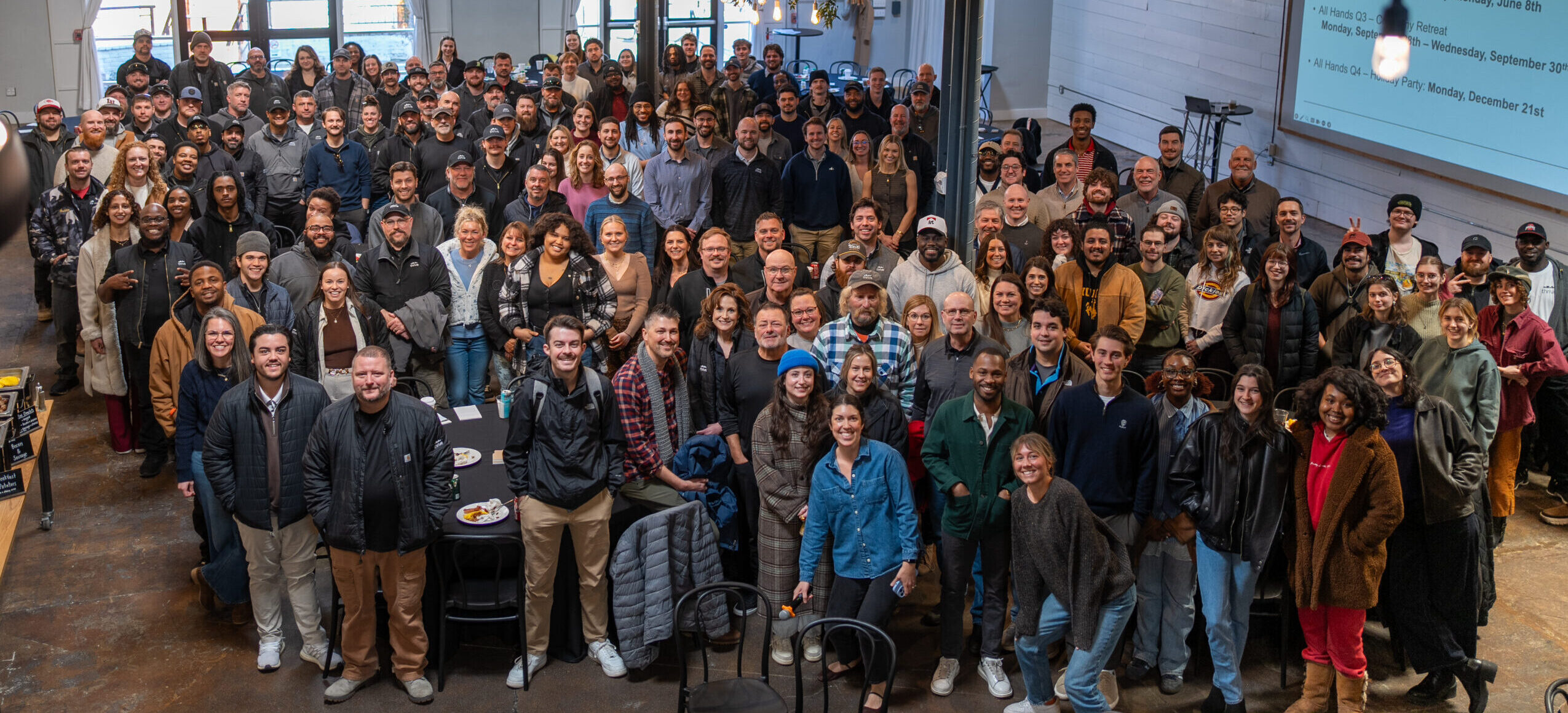 Thrive Companies, Our Team gathers for a group photo inside a spacious, modern room with exposed ceilings and a presentation screen on the wall. Apartments in Columbus Ohio