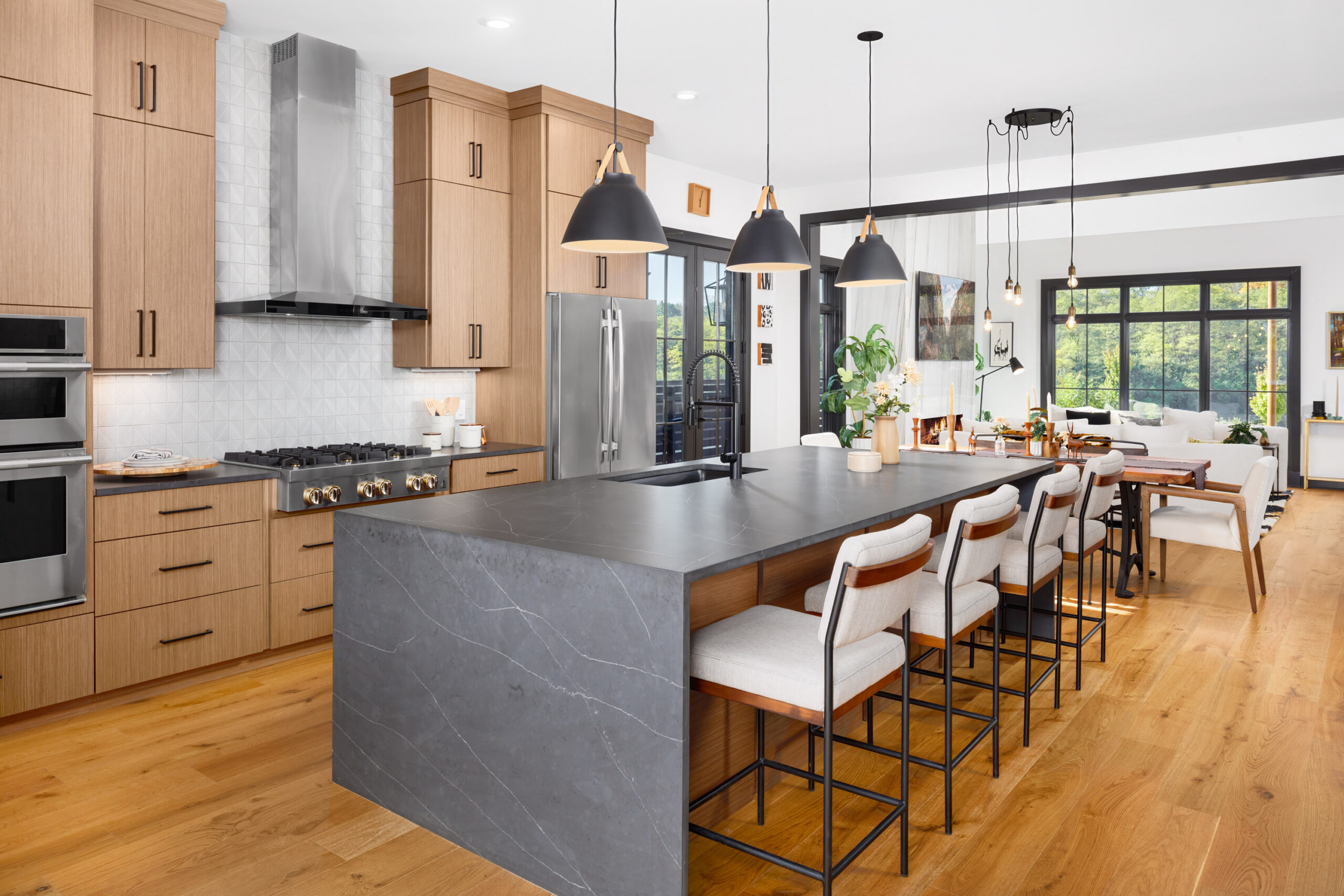 Thrive Companies, Modern kitchen with wood cabinets, black countertops, a large island with barstools, pendant lights, and a dining area leading to a bright living room with large windows. Apartments in Columbus Ohio