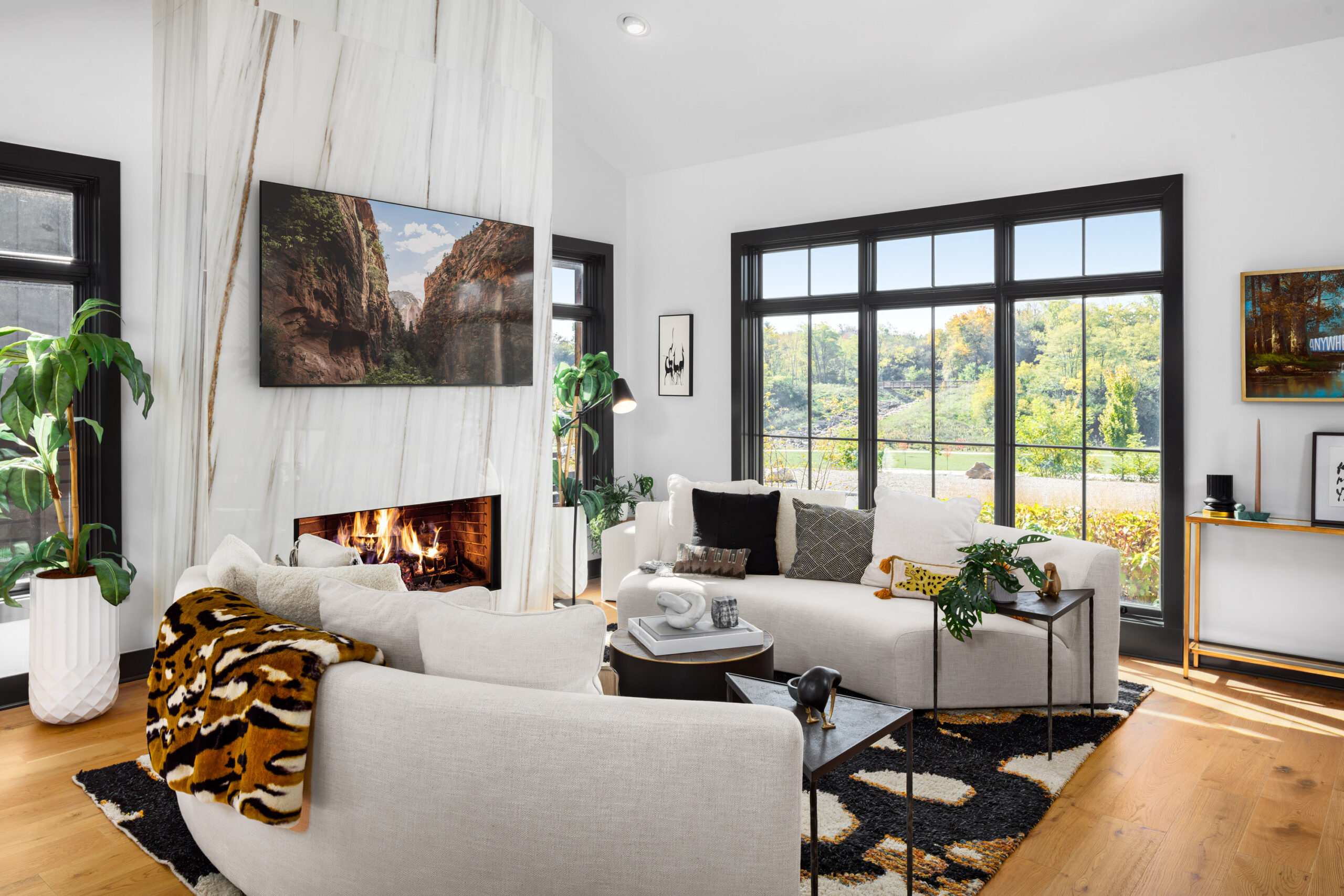 Thrive Companies, Modern living room with white sofas, a patterned rug, indoor plants, a marble fireplace with a TV above, large windows, and outdoor greenery visible through the glass. Apartments in Columbus Ohio