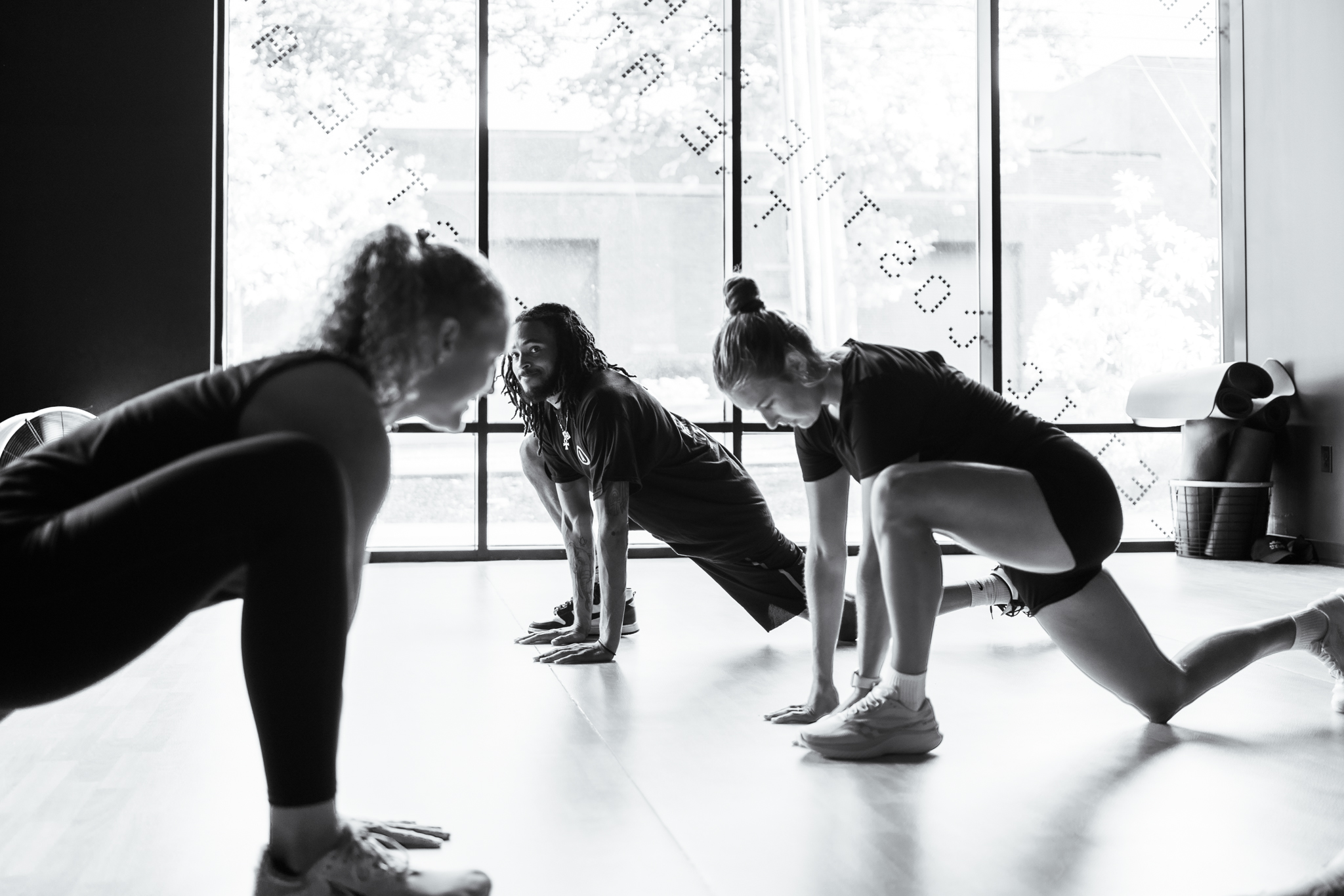 Thrive Companies, Three people in athletic wear perform lunging stretches on the floor of a fitness studio with large windows in the background. Apartments in Columbus Ohio