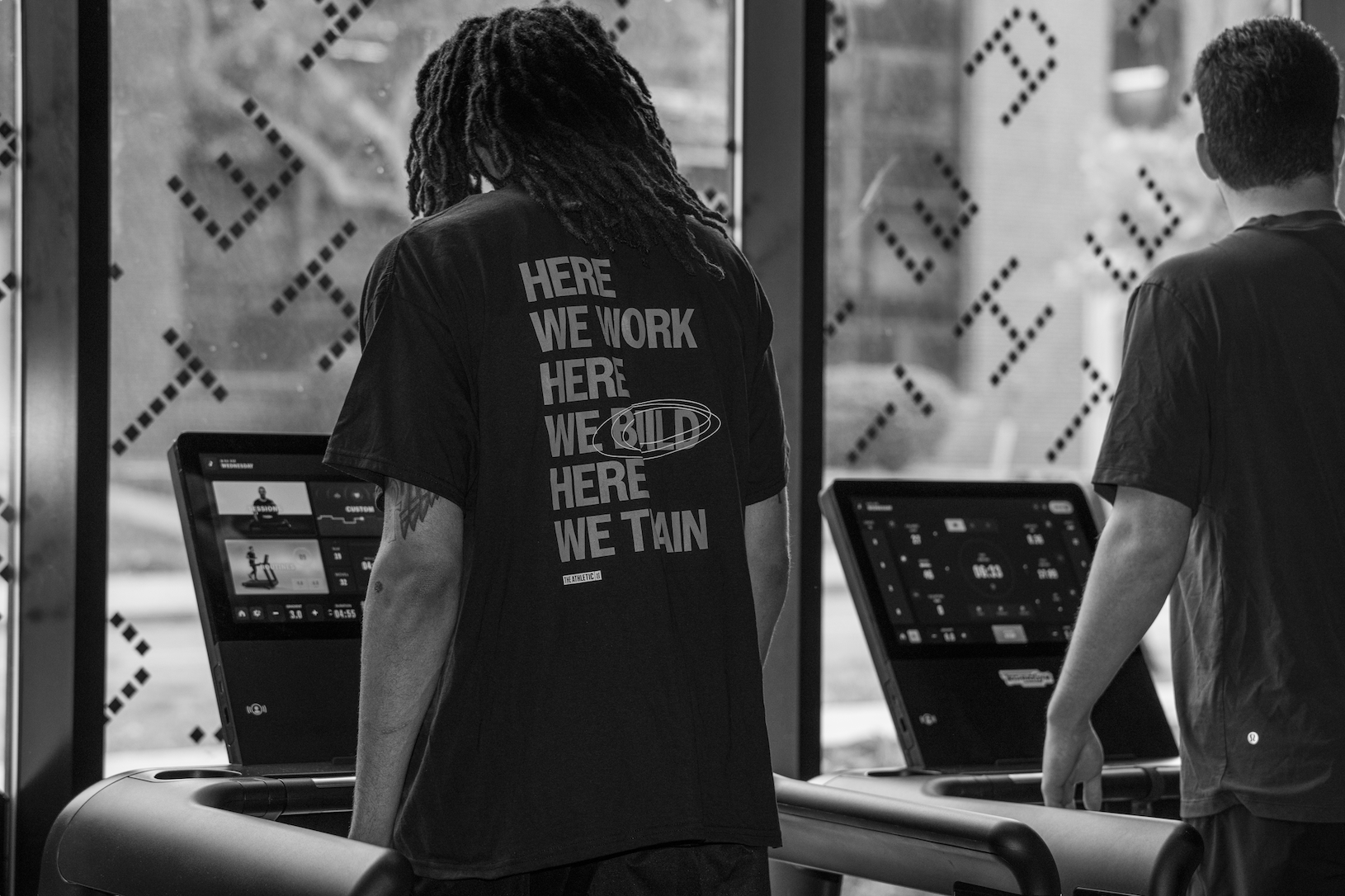Thrive Companies, Two people stand on treadmills in a gym. One wears a shirt that says, HERE WE WORK HERE WE BUILD HERE WE TRAIN. The image is in black and white. Apartments in Columbus Ohio