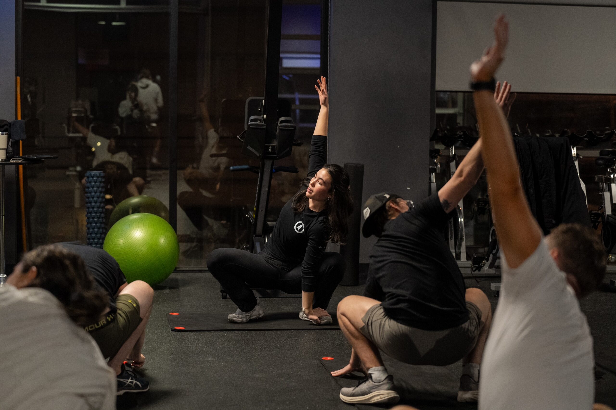 Thrive Companies, A fitness instructor leads a group stretching session indoors, with participants mirroring her raised arm pose on exercise mats. Gym equipment and a green exercise ball are visible in the background. Apartments in Columbus Ohio