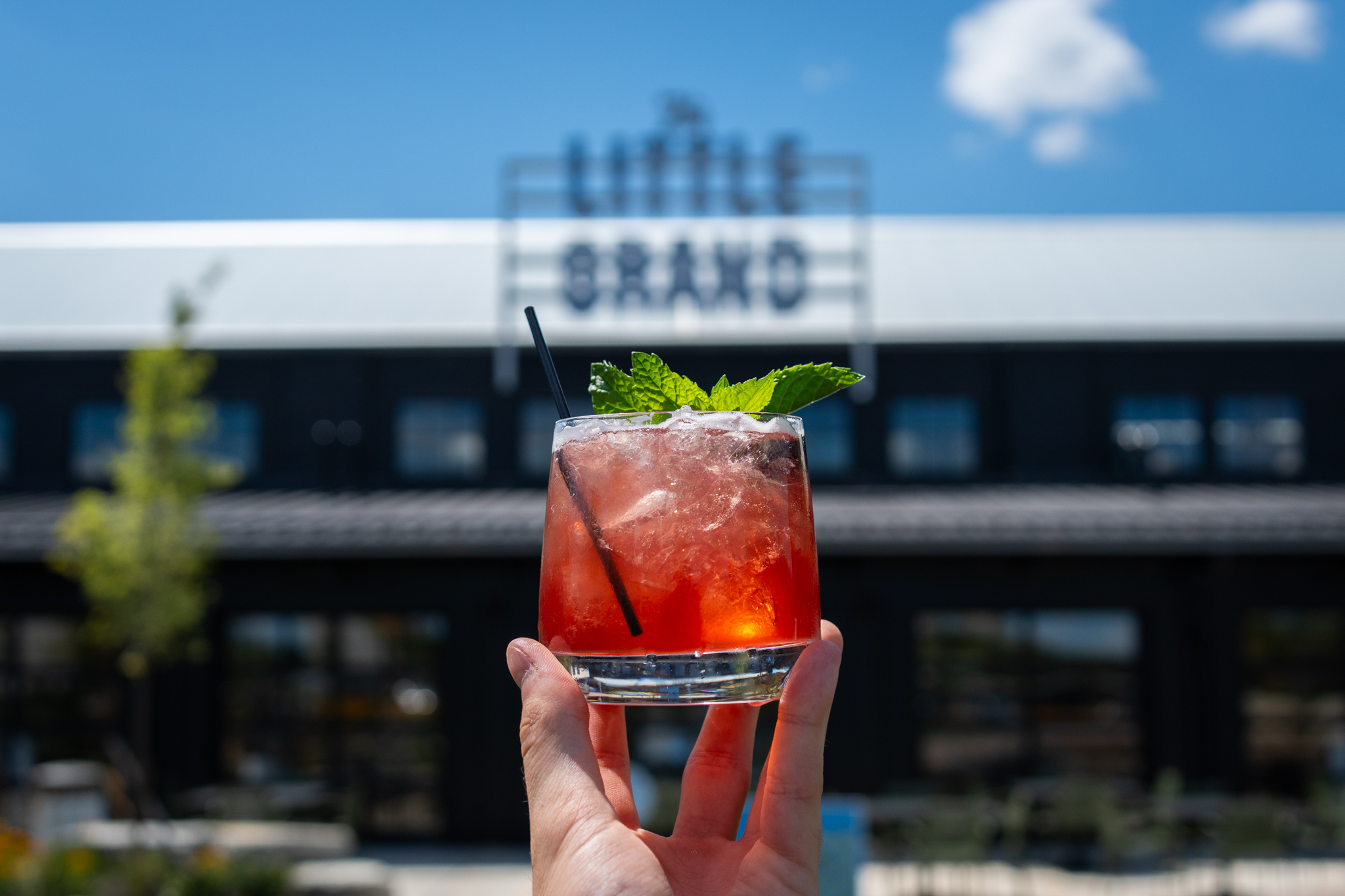 Thrive Companies, A hand holds a glass of red cocktail with mint leaves and a black straw, with a blurred building by Thrive Companies and blue sky in the background. Apartments in Columbus Ohio