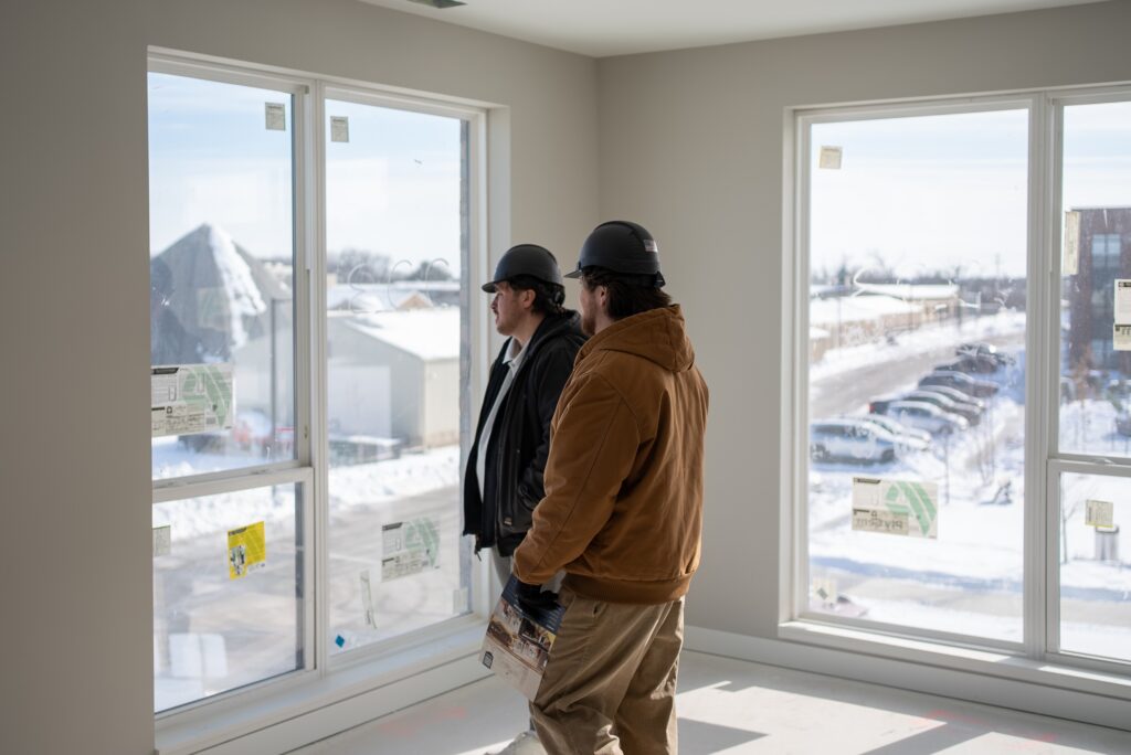 Thrive Companies, Two construction workers wearing hard hats stand inside a newly built construction site with large windows, looking out at a snowy landscape and parked cars. Apartments in Columbus Ohio