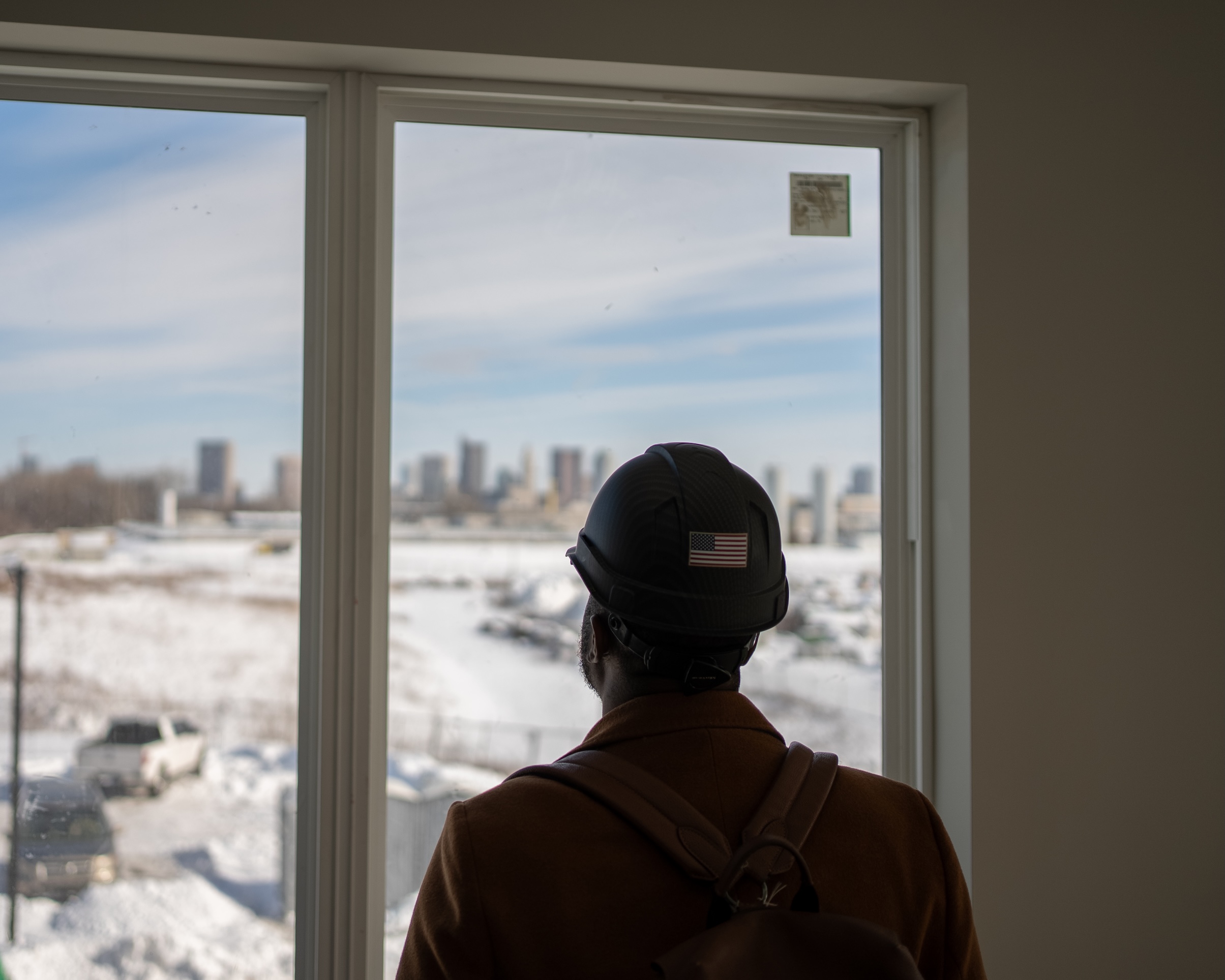Thrive Companies, A person wearing a black construction hard hat and brown coat looks out a window at a snow-covered landscape and distant city skyline. Apartments in Columbus Ohio