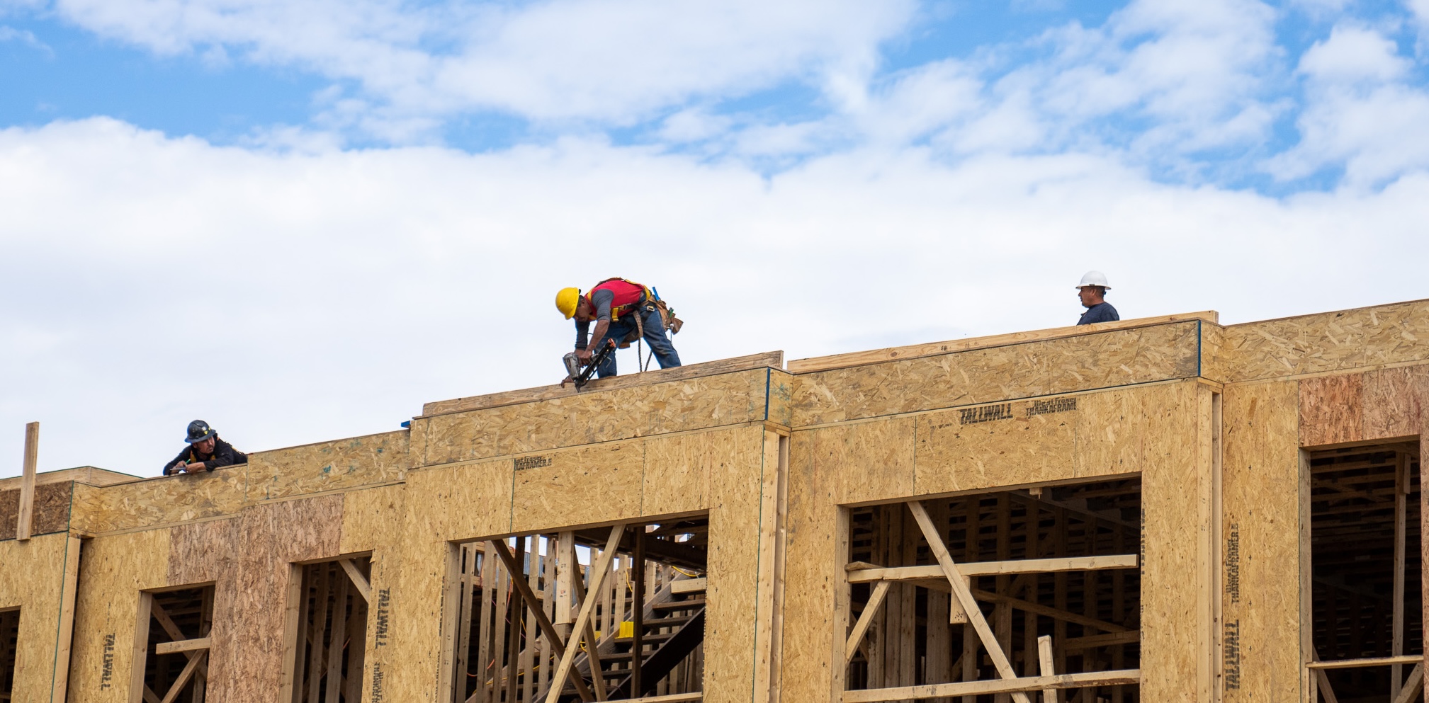 Thrive Companies, Three construction workers in safety gear work on the wooden frame of a multi-story construction project under a blue sky with clouds. Apartments in Columbus Ohio