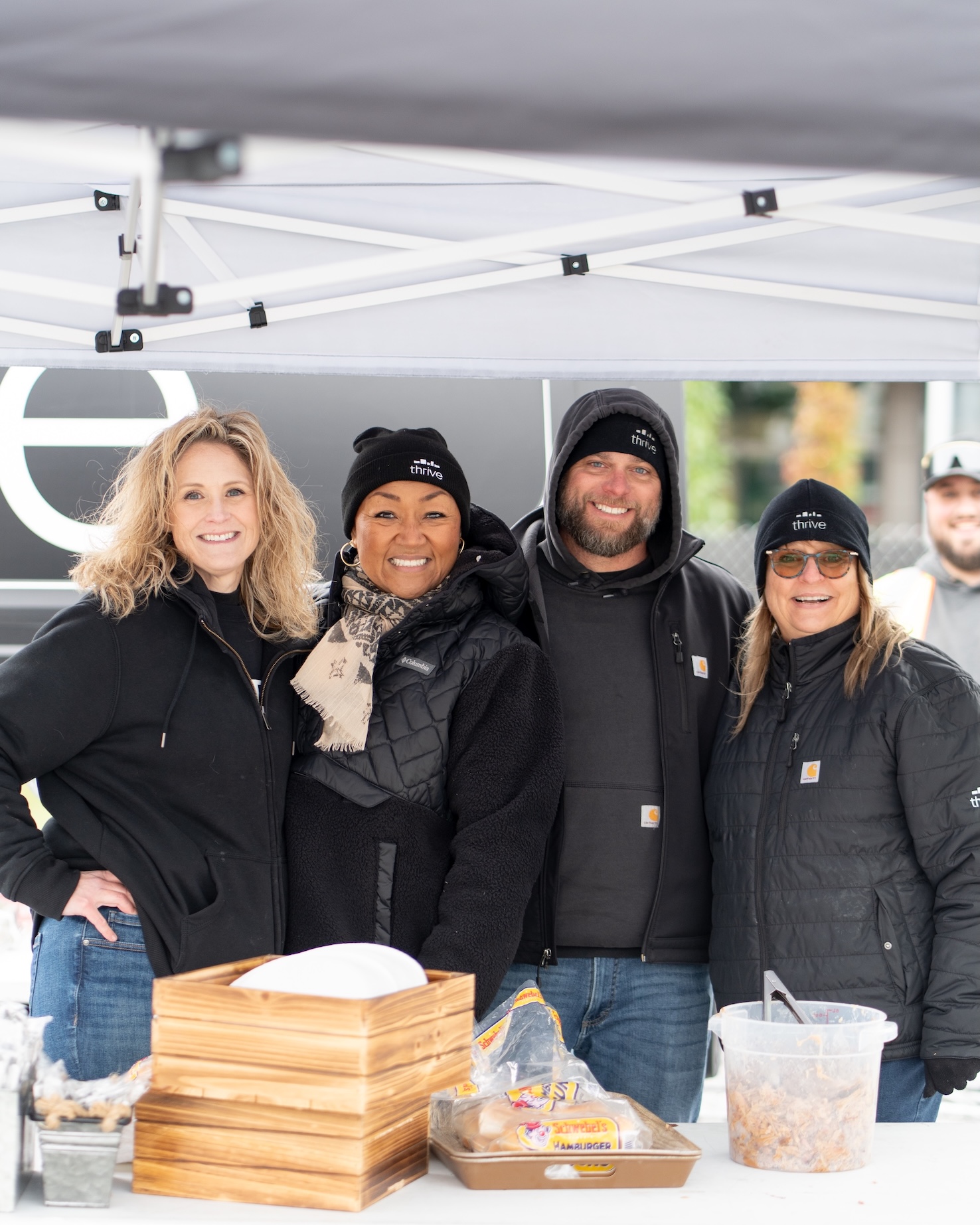 Thrive Companies, Four people wearing black jackets and beanies stand together and smile under a canopy at an outdoor event, with food containers on the table in front of them. Apartments in Columbus Ohio