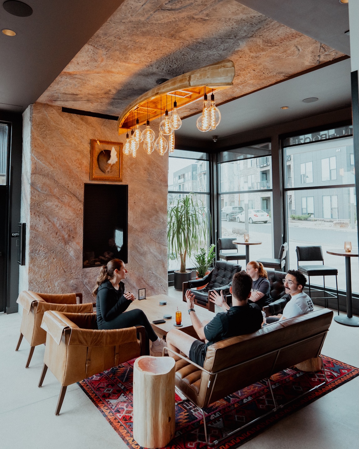 Thrive Companies, Four people sit and talk in a modern lounge area by Thrive Companies, featuring leather chairs, large windows, and a hanging light fixture. Apartments in Columbus Ohio