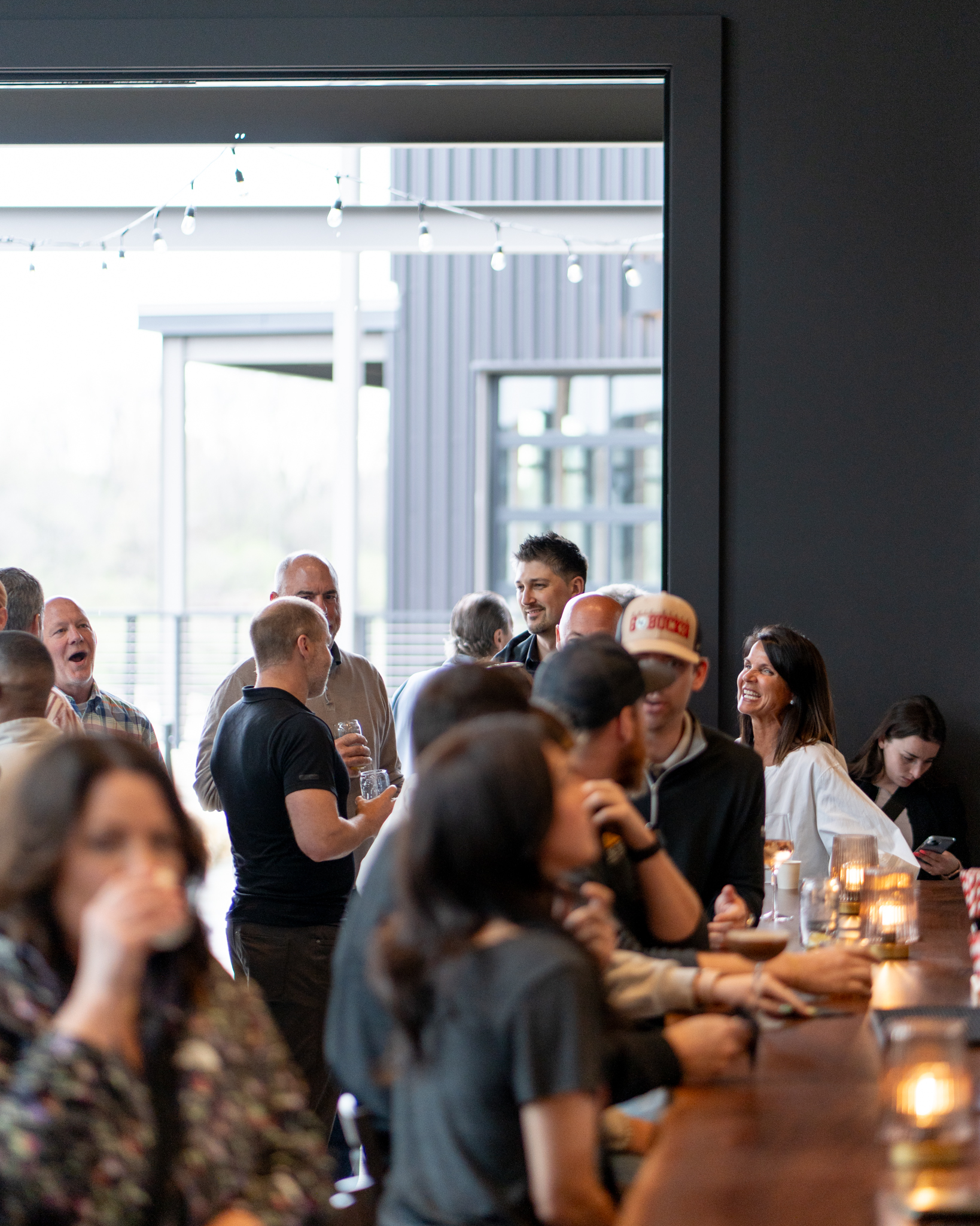 Thrive Companies, A group of people socialize and drink at a modern indoor bar, with large windows showing an outdoor patio in the background. Apartments in Columbus Ohio