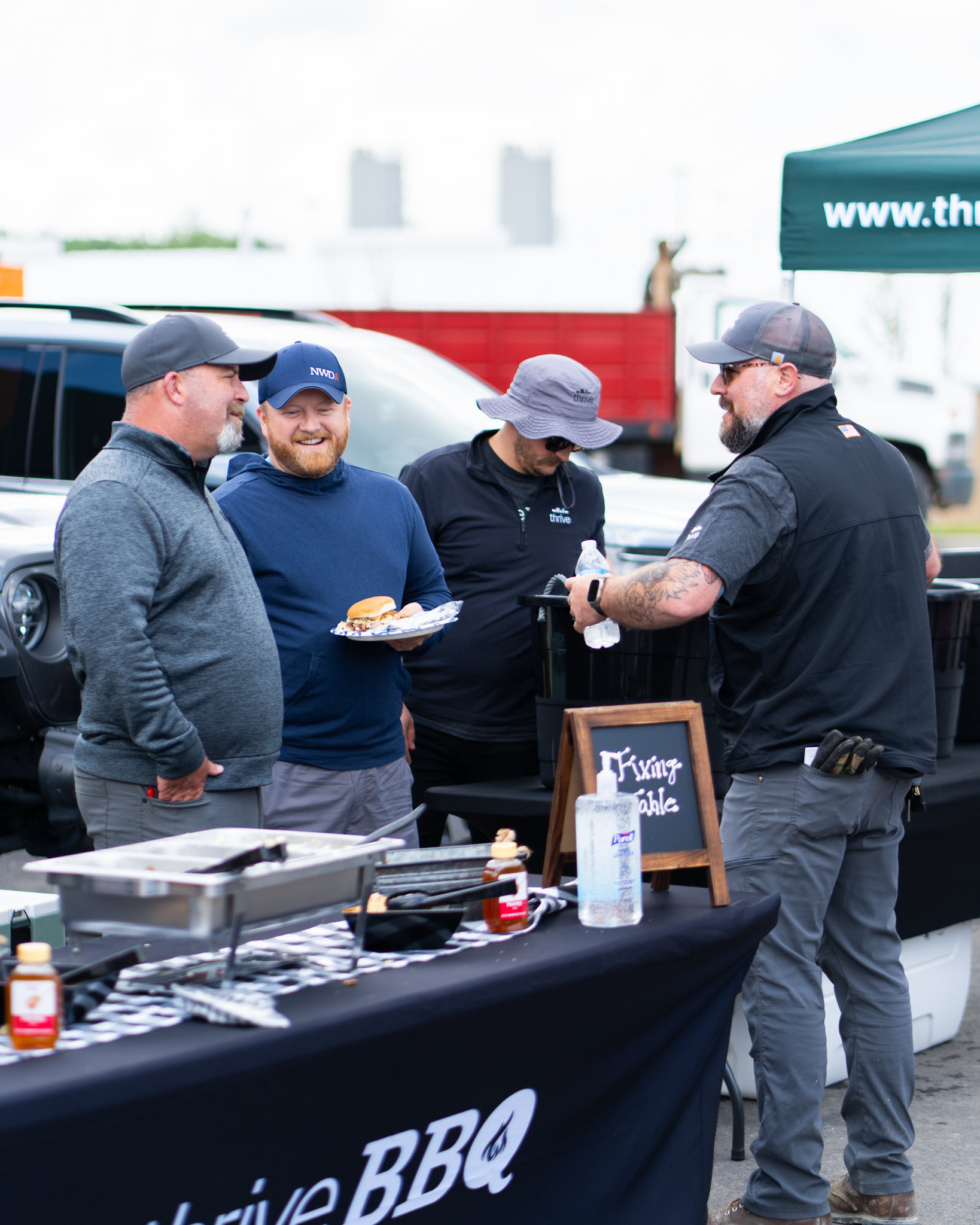Thrive Companies, Four men stand and talk near a BBQ setup with food trays, condiments, and a sign that reads Thrive BBQ at an outdoor event. Apartments in Columbus Ohio