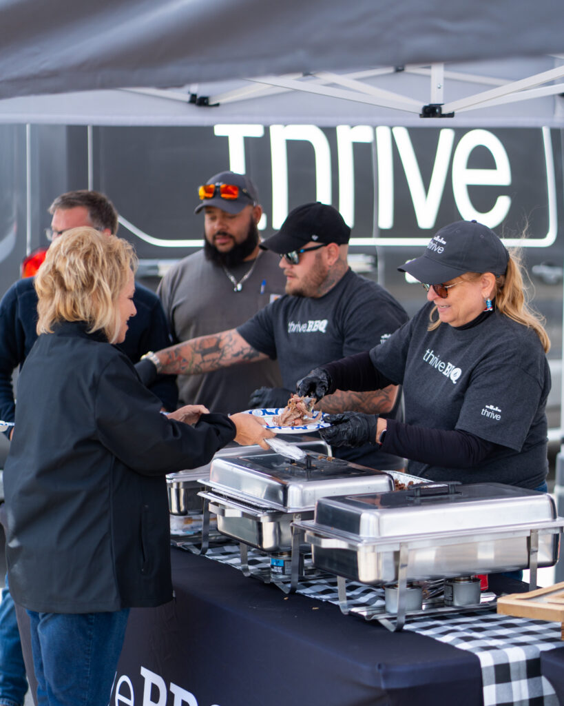 Thrive Companies, People serve food to guests from buffet trays at an outdoor event under a canopy, with the Thrive Companies Ohio logo visible in the background. Apartments in Columbus Ohio