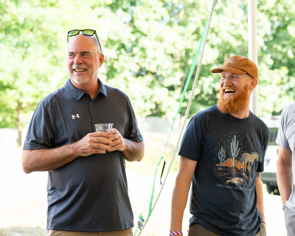Thrive Companies, Two men standing outdoors, smiling and talking; one holds a drink, both are casually dressed with sunglasses, enjoying the day in a Thrive Companies Ohio community, with trees blurred in the background. Apartments in Columbus Ohio