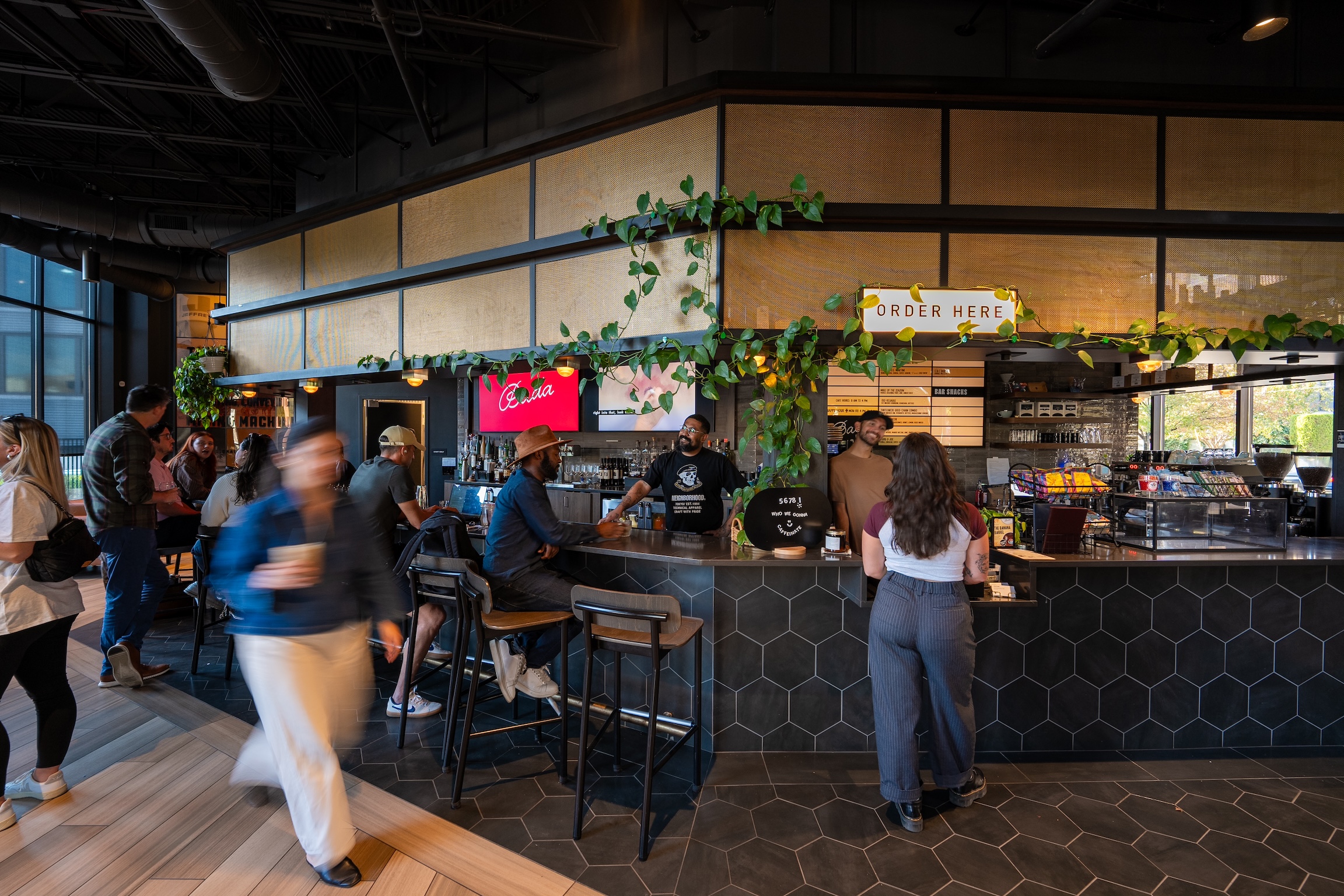 Thrive Companies, People order and socialize at a modern coffee shop counter with black hexagonal tiles, hanging plants, and a sign reading Order Here, while our team ensures every visit feels welcoming and memorable. Apartments in Columbus Ohio