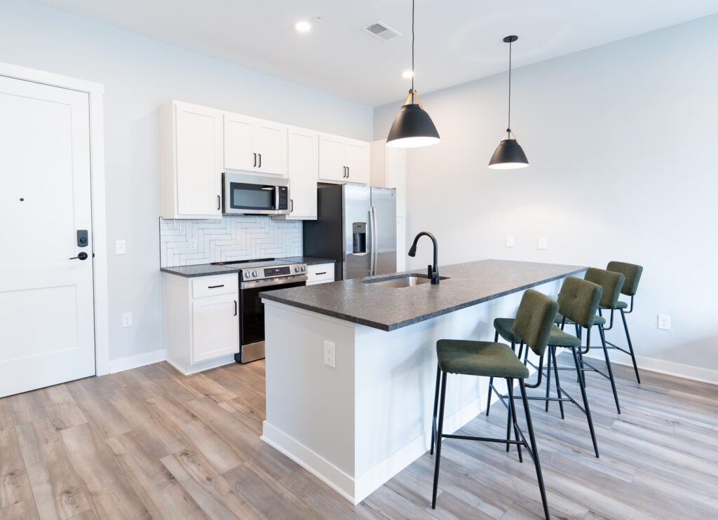 Thrive Companies, Modern kitchen with white cabinets, stainless steel appliances, a black countertop island with sink, and three green barstools&mdash;a stylish space perfect for gatherings or community management meetings. Two pendant lights hang above the island. Apartments in Columbus Ohio