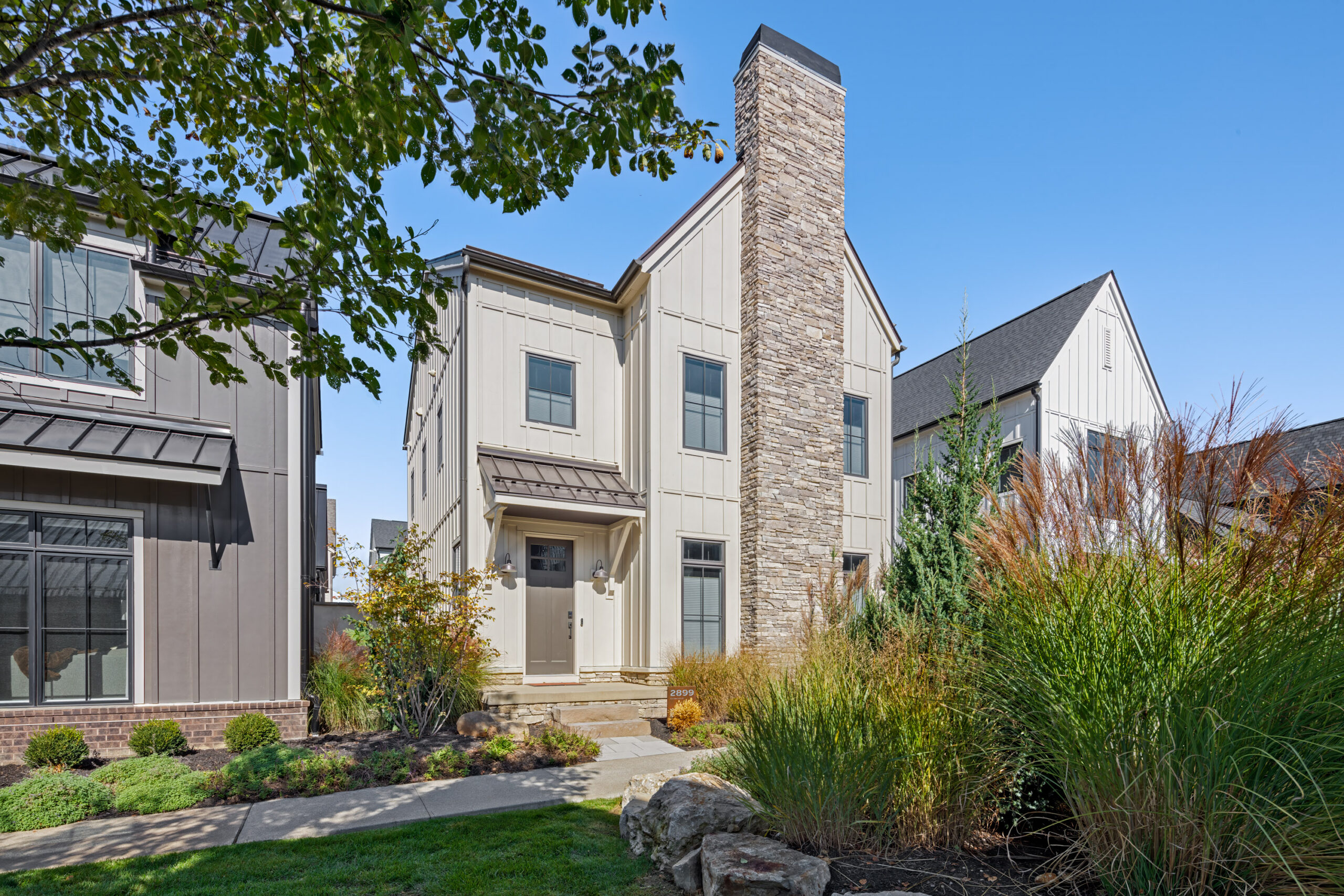 Thrive Companies, A modern two-story house with vertical siding, a prominent stone chimney, and landscaped front yard, set among similar neighboring homes under a clear blue sky. Apartments in Columbus Ohio