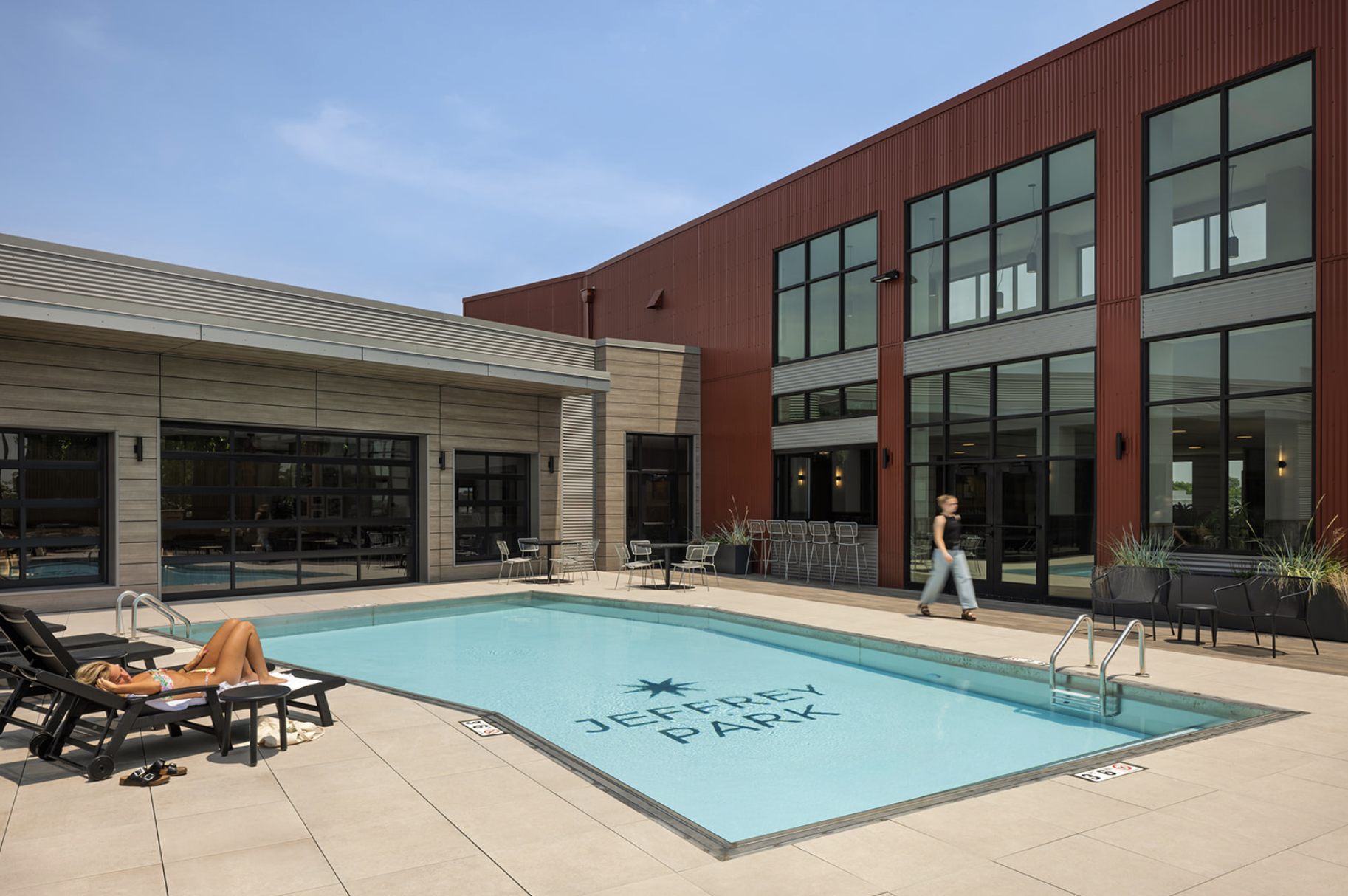 Thrive Companies, Outdoor pool area at a modern apartment complex with lounge chairs, a person sunbathing, another walking, and large windows in the background. Ask about our Resident Referral Program while enjoying Jeffrey Park by the pool. Apartments in Columbus Ohio