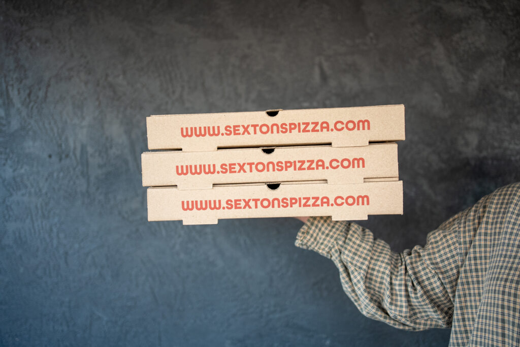 Thrive Companies, A person in a plaid shirt holds three stacked pizza boxes labeled www.sextonspizza.com, spotlighting the tasty offerings from Sexton’s Pizza at Little Grand Market, against a dark textured background. Apartments in Columbus Ohio