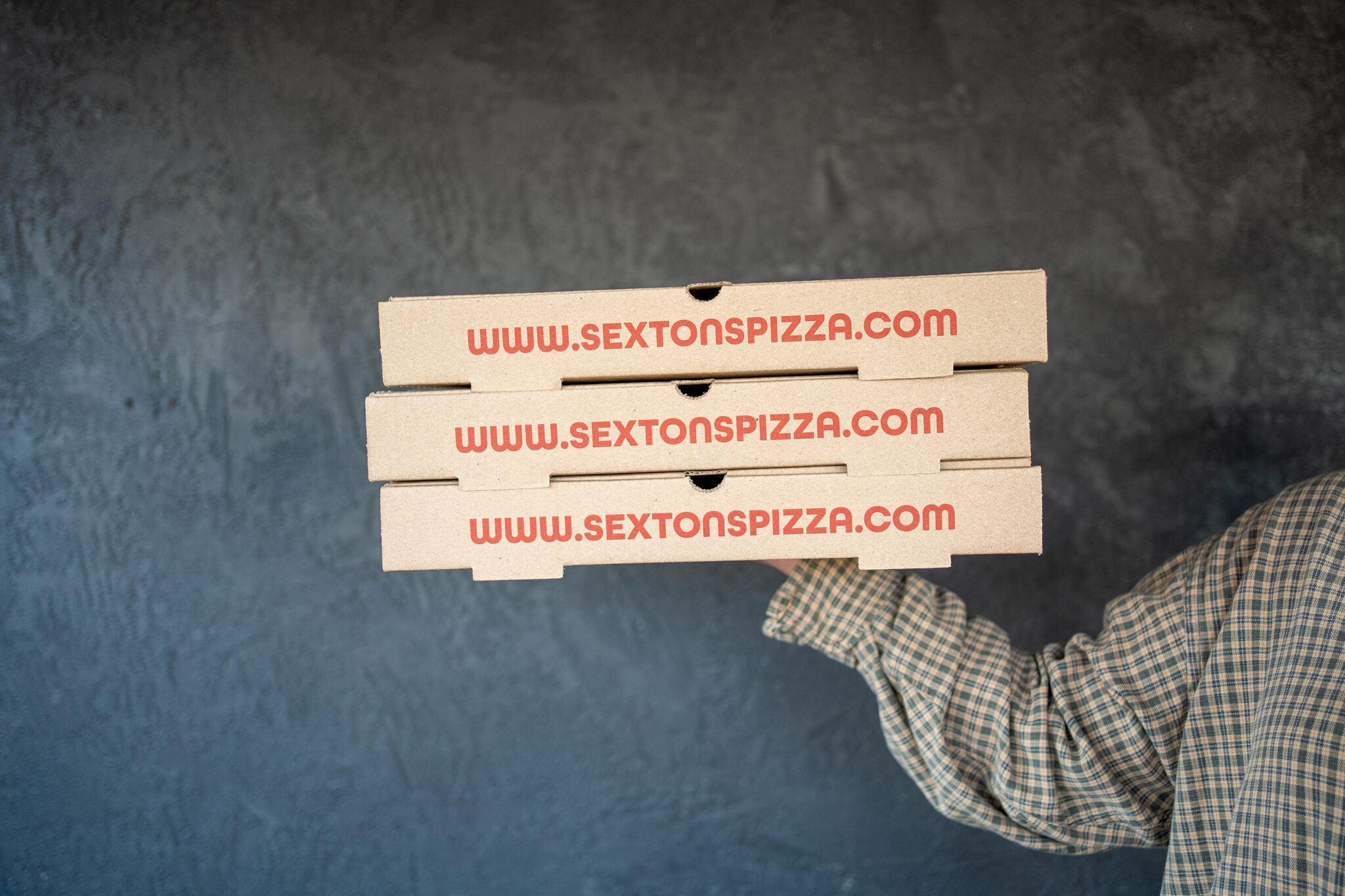 Thrive Companies, A person in a plaid shirt holds three stacked pizza boxes labeled www.sextonspizza.com, spotlighting the tasty offerings from Sexton’s Pizza at Little Grand Market, against a dark textured background. Apartments in Columbus Ohio