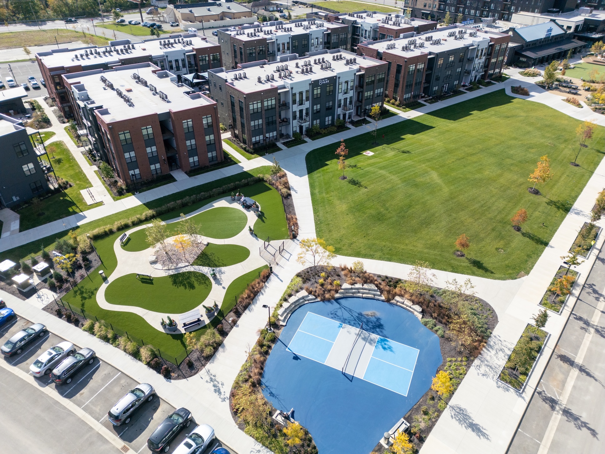 Thrive Companies, Aerial view of a modern apartment complex featuring a large grassy courtyard, walking paths, a playground area, and a small blue basketball court surrounded by parked cars. Apartments in Columbus Ohio