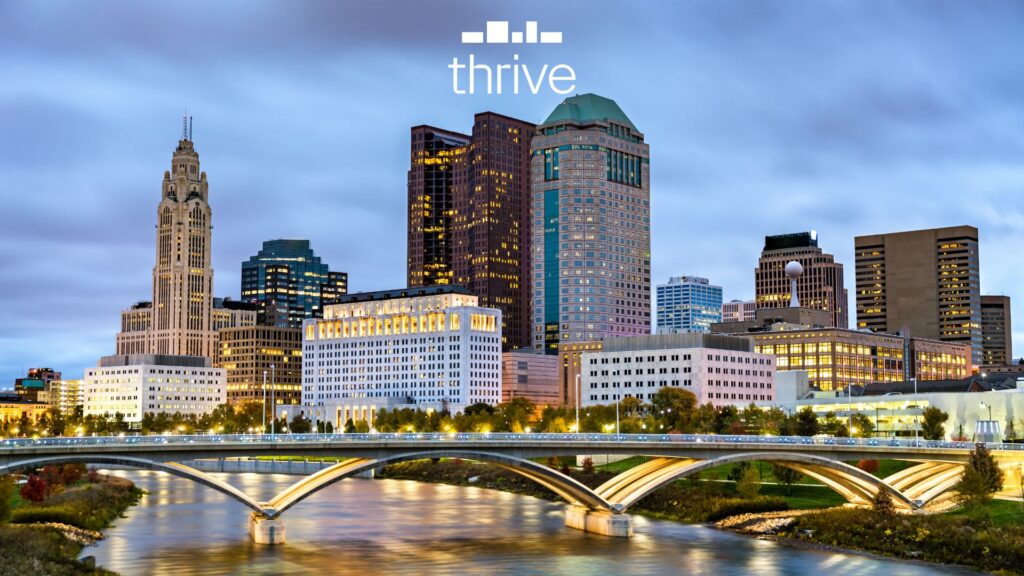 Thrive Companies, A city skyline at dusk with modern and historic buildings, a river and bridge in the foreground, set in the vibrant Franklinton Arts District, with the word thrive at the top. Apartments in Columbus Ohio